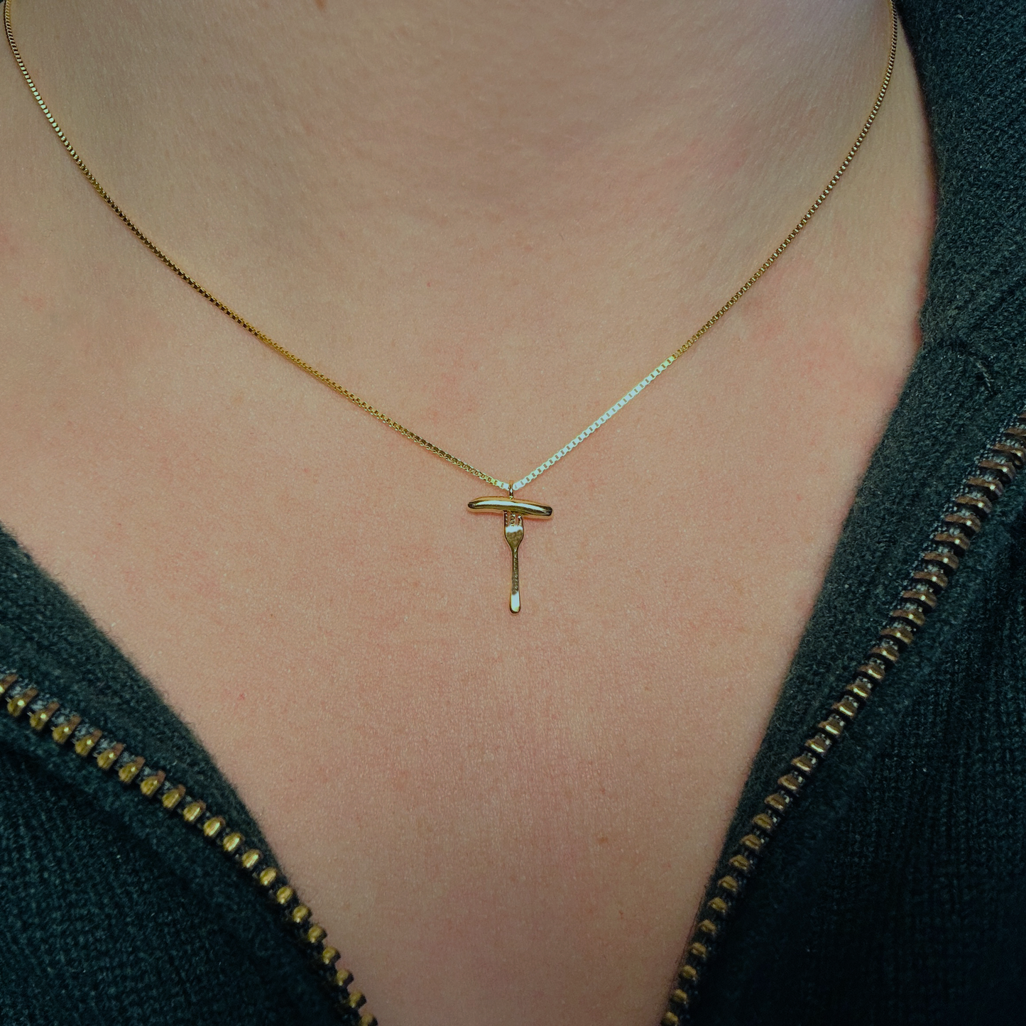 Eine goldene Halskette mit einem filigranen Anhänger in Form einer Bratwurst auf einer Gabel wird um den Hals einer Person getragen. Die feine Kette mit 3 Micron Vergoldung auf Sterling Silber (925) liegt dezent auf der Haut und verleiht dem Schmuckstück eine elegante Ausstrahlung. Der Anhänger zeigt eine stilisierte Bratwurst auf einer Gabel, die für Tradition und Genuss steht. Die Person trägt eine dunkelgrüne Jacke mit goldenem Reißverschluss, die den edlen Charakter des Schmucks unterstreicht.