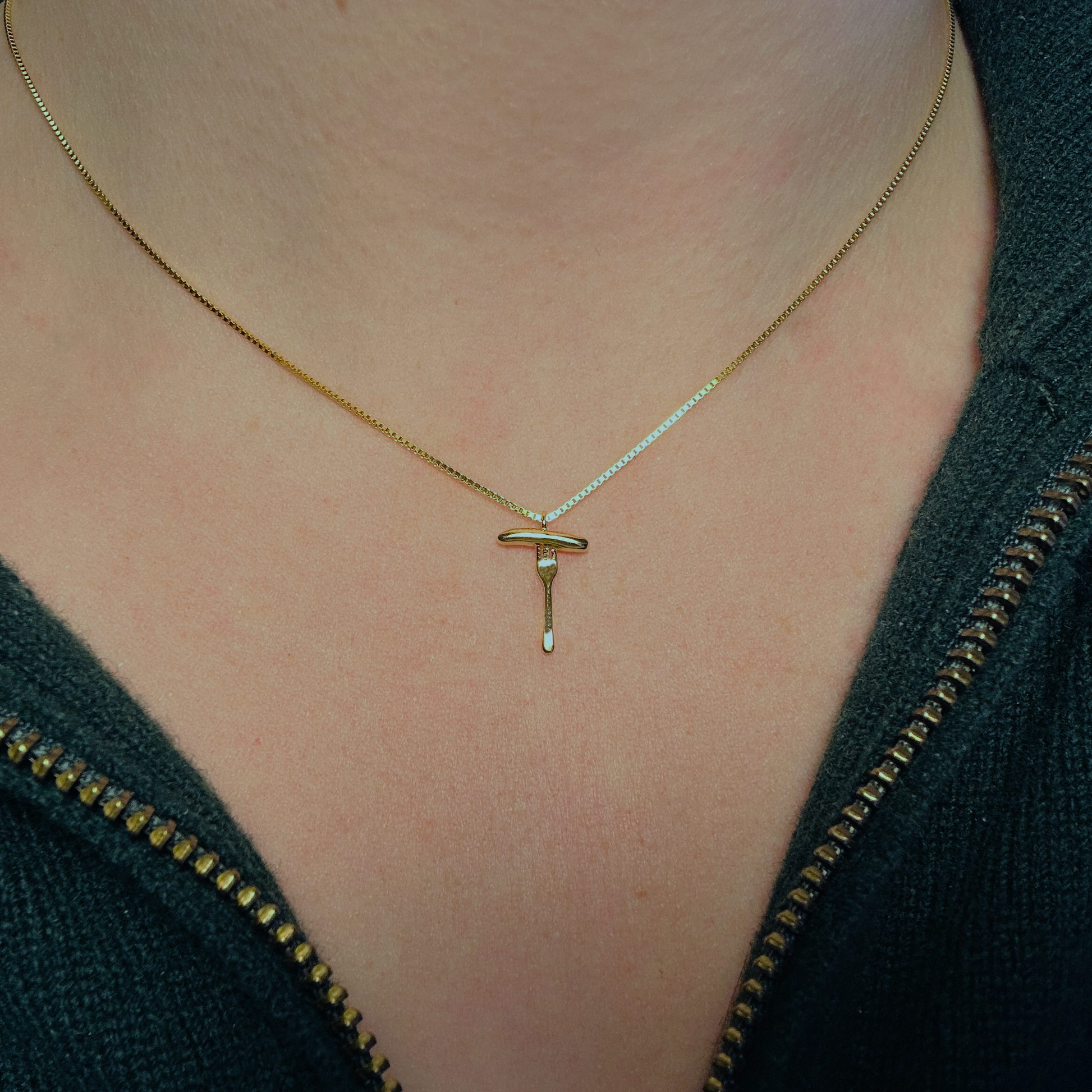Eine goldene Halskette mit einem filigranen Anhänger in Form einer Bratwurst auf einer Gabel wird um den Hals einer Person getragen. Die feine Kette mit 3 Micron Vergoldung auf Sterling Silber (925) liegt dezent auf der Haut und verleiht dem Schmuckstück eine elegante Ausstrahlung. Der Anhänger zeigt eine stilisierte Bratwurst auf einer Gabel, die für Tradition und Genuss steht. Die Person trägt eine dunkelgrüne Jacke mit goldenem Reißverschluss, die den edlen Charakter des Schmucks unterstreicht.