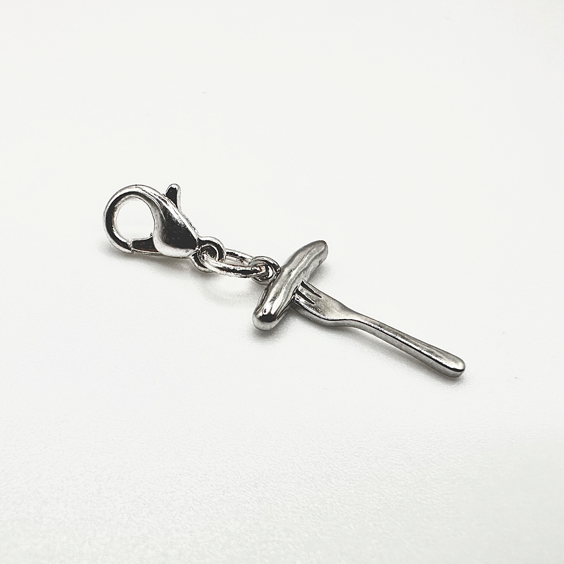 Nahaufnahme eines silbernen Anhängers in Form einer Bratwurst auf einer Gabel mit einem Karabinerhaken als Verschluss. Der detaillierte Charm besteht aus echtem Sterling Silber (925) und ist durch seine glänzende Oberfläche ein stilvoller Blickfang. Dank des praktischen Verschlusses lässt sich der Anhänger leicht an Ketten oder Armbändern befestigen. Ideal für Liebhaber fränkischer Kulinarik und originellen Schmucks.