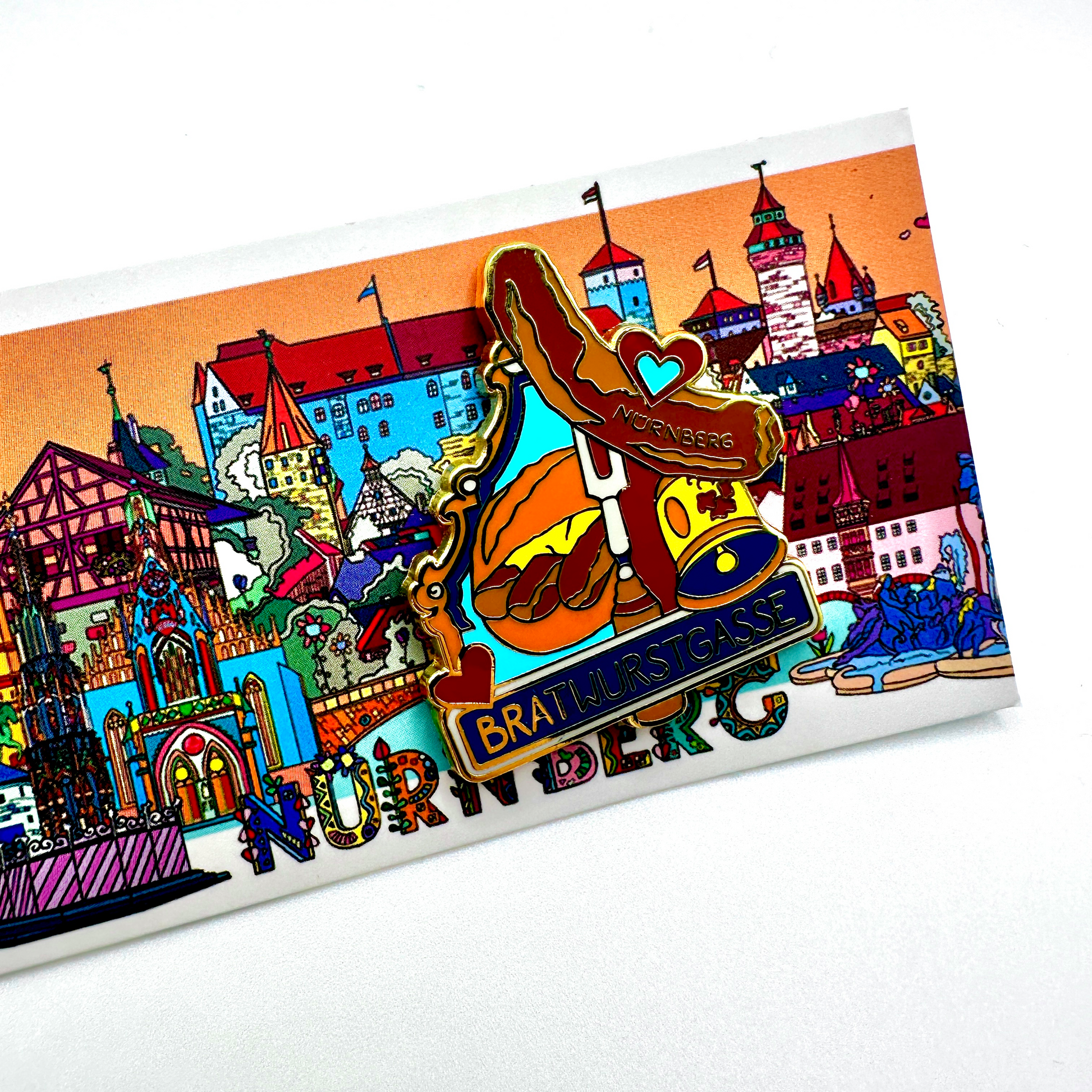 Bunter Emaille-Pin mit der Aufschrift „Bratwurstgasse“ und einem detailreichen Design: eine Bratwurst auf einer Gabel mit einem Herz und dem Schriftzug „Nürnberg“, sowie ein traditionelles „3 im Weggla“-Brötchen. Im Hintergrund eine farbenfrohe Illustration der Nürnberger Altstadt mit Wahrzeichen wie der Kaiserburg und der Frauenkirche. Ein originelles Andenken für Liebhaber fränkischer Bratwurstkultur.