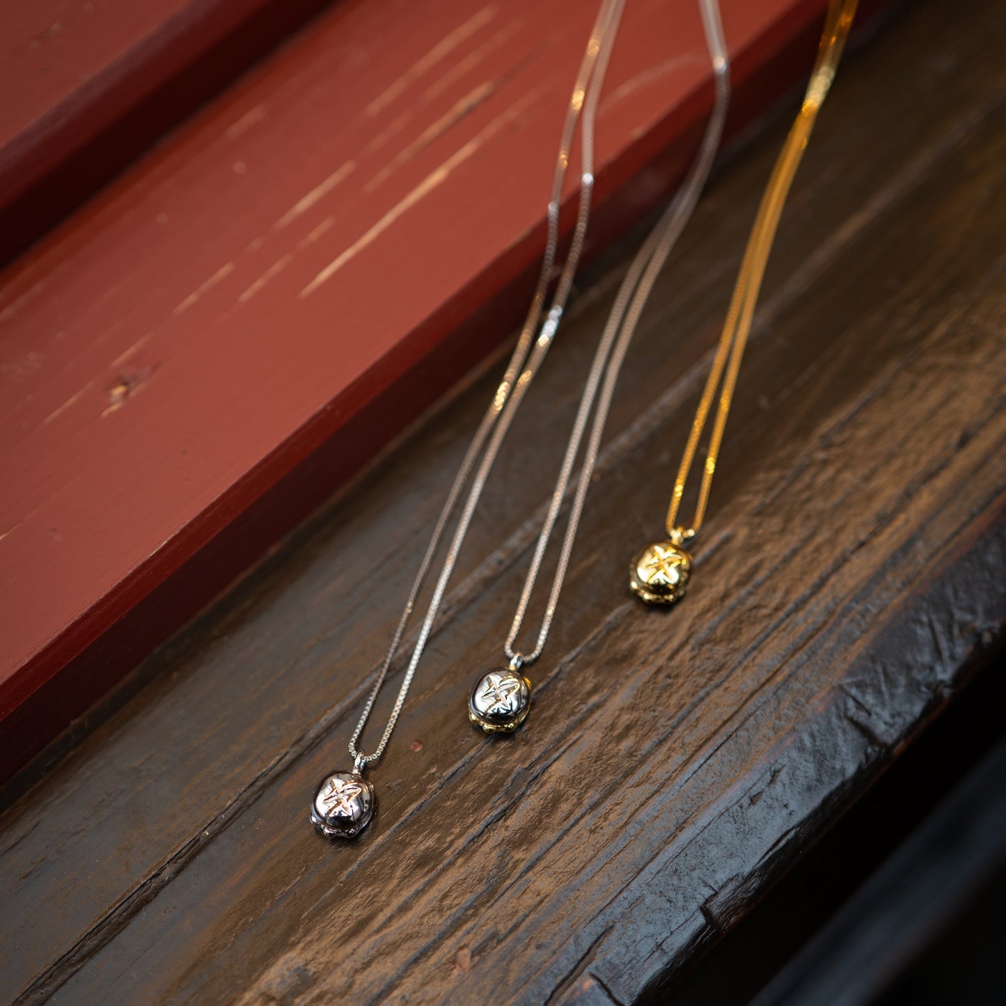 Drei filigrane Ketten mit kleinen, runden Anhängern liegen dekorativ auf einer dunklen Holzoberfläche. Die Anhänger sind in Form eines Brötchens mit einer charakteristischen Prägung gestaltet. Zwei der Ketten bestehen aus Sterling-Silber (925), während die dritte mit einer hochwertigen Vergoldung veredelt ist. Die Kombination aus Silber- und Goldtönen verleiht dem Schmuckstück eine edle und elegante Ausstrahlung. Im Hintergrund ist eine rötlich-braune Holzstruktur zu sehen.