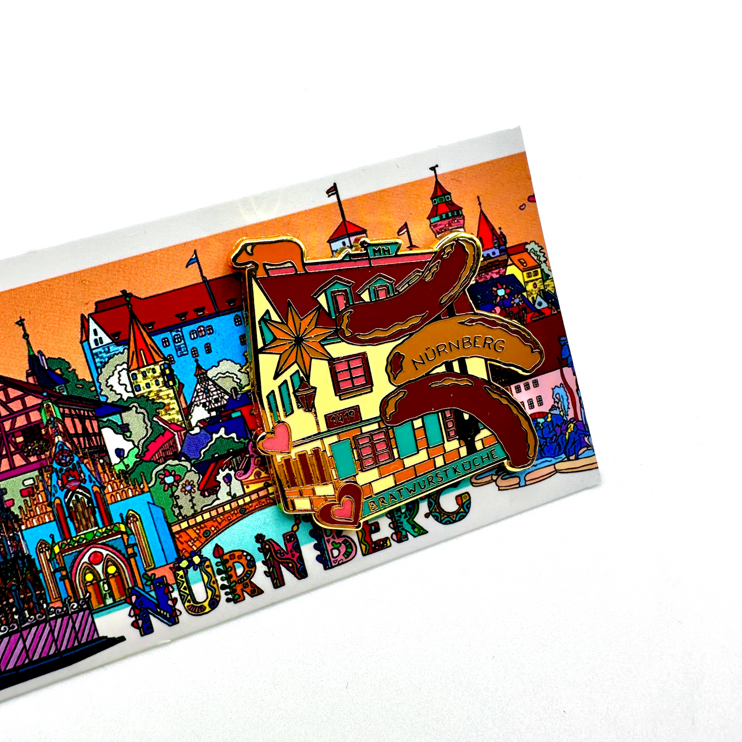 Detailreicher Emaille-Pin mit einer Darstellung der historischen Nürnberger Bratwurstküche. Das Motiv zeigt das Fachwerkhaus der ältesten Bratwurstküche der Welt mit goldenen Akzenten, zwei Herzen und drei Bratwürsten mit der Aufschrift „Nürnberg“. Im Hintergrund eine farbenfrohe Illustration der Nürnberger Altstadt mit Wahrzeichen wie der Kaiserburg und der Frauenkirche. Ein einzigartiges Souvenir für Liebhaber der fränkischen Bratwursttradition.