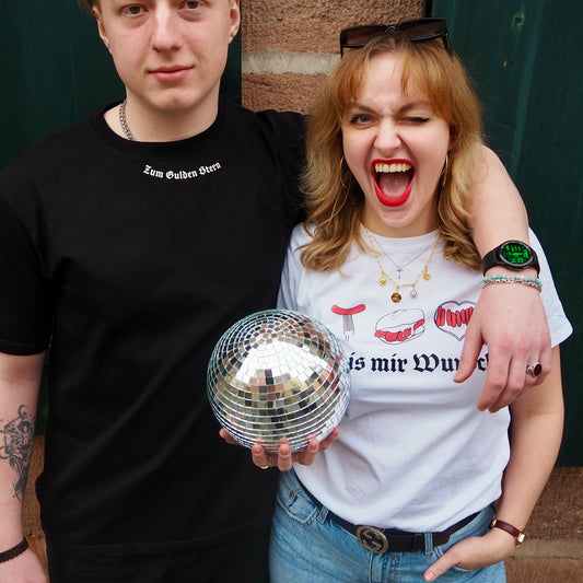 Ein junger Mann und eine junge Frau posieren gemeinsam vor einer rustikalen Steinmauer. Der Mann trägt ein schwarzes T-Shirt mit der Aufschrift "Zum Gulden Stern", während die Frau ein weißes T-Shirt mit Bratwurst-Illustrationen und dem Text "is mir Wurst" trägt. Sie hält eine Discokugel in der Hand und lacht mit einem Augenzwinkern in die Kamera. Beide tragen Schmuck und Accessoires, die ihren individuellen Stil unterstreichen. Die Szene strahlt eine lockere und fröhliche Stimmung aus.