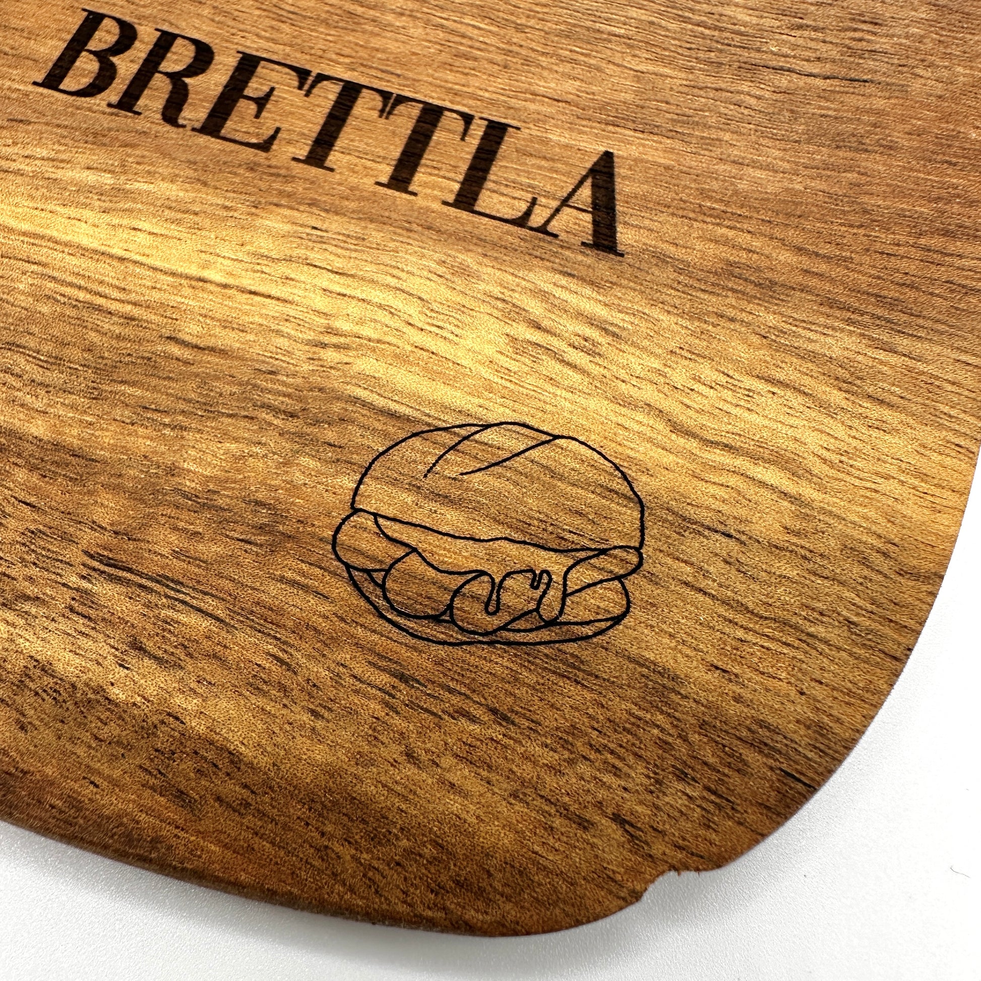 Detailaufnahme eines hochwertigen Holzbretts mit der Gravur "BRETTLA" in schwarzer Schrift. In der unteren rechten Ecke ist eine feine Gravur eines Brötchens mit drei Bratwürsten zu sehen – ein stilisiertes Symbol für die fränkische Spezialität "3 im Weggla". Die natürliche Holzmaserung verleiht dem Brett eine rustikale und edle Optik.