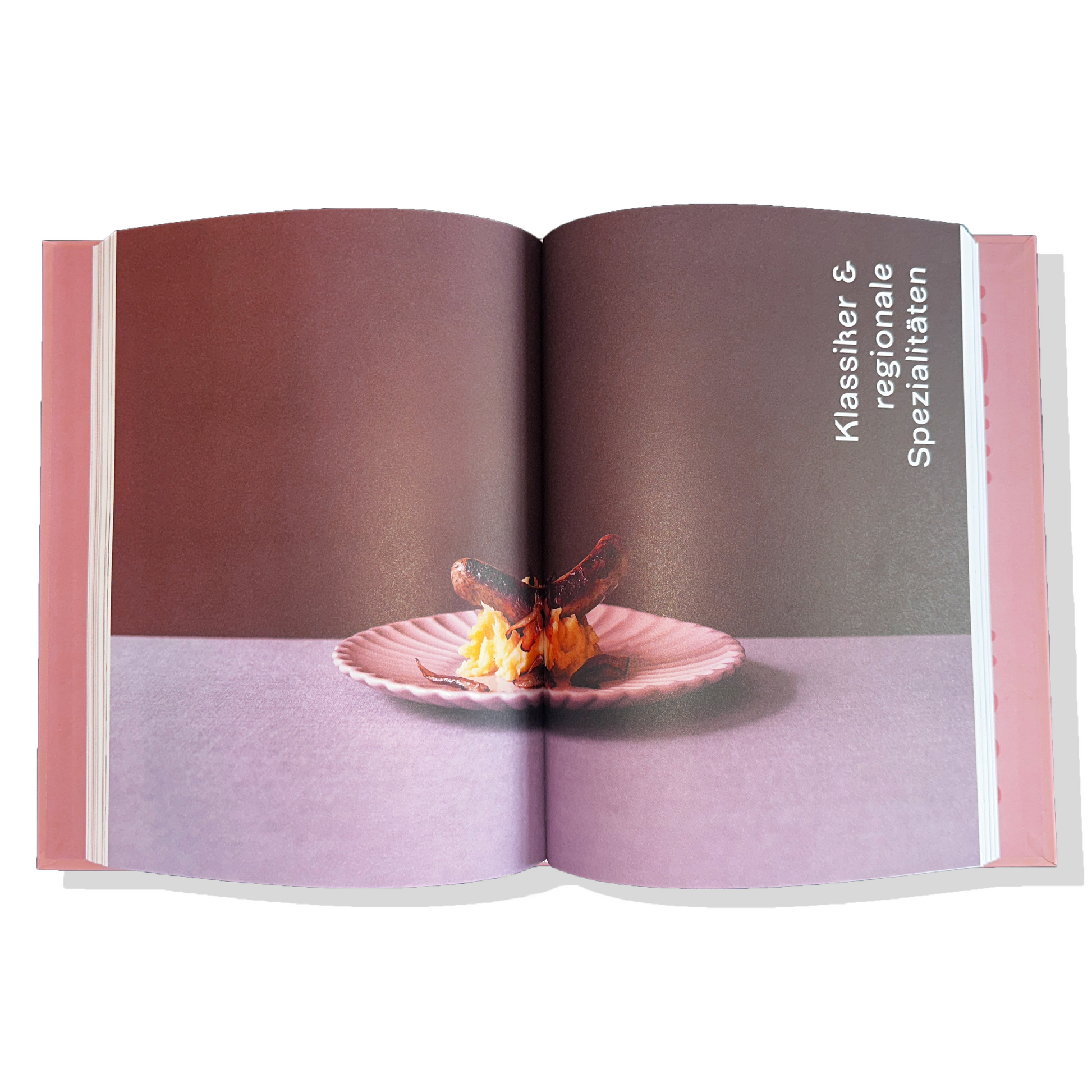 "Ein aufgeschlagenes Buch mit einer Food-Fotografie auf einer Doppelseite. Auf einem rosa Muschelteller liegt ein kunstvoll angerichtetes Gericht mit einer dunklen Wurst, die auf einem Bett aus Kartoffelpüree stehen. Der Hintergrund ist dunkelbraun, während die Tischoberfläche in einem sanften Lila-Ton gehalten ist. Auf der rechten Seite des Buches steht in weißer Schrift der Text 'Klassiker & regionale Spezialitäten' vertikal geschrieben."