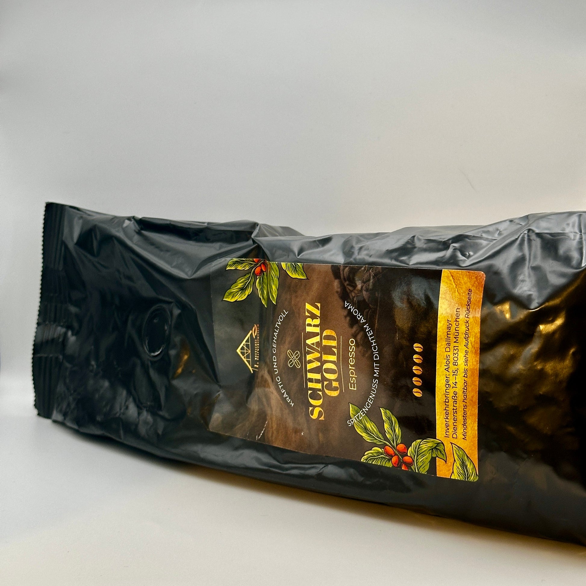 Eine edle, schwarze Kaffeeverpackung mit goldener Aufschrift „Schwarz Gold – Espresso“ liegt seitlich. Das Design ist mit Kaffeepflanzen-Illustrationen verziert und betont den hochwertigen, kräftigen und gehaltvollen Geschmack des Espressos. Die Verpackung stammt von Dallmayr und verspricht einen intensiven Kaffeegenuss mit dichtem Aroma.