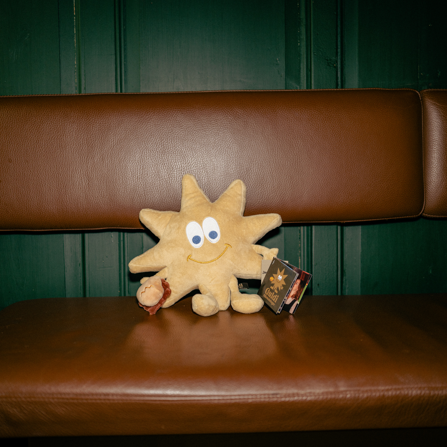 Ein weiches, sternförmiges Kuscheltier mit lächelndem Gesicht sitzt auf einer braunen Lederbank vor einer grünen Holzwand. Guldi, dein Glücksbringer, hält 3 im Weggla in der Hand und trägt ein Etikett mit seinem Namen. Die Szene vermittelt eine warme, gemütliche Atmosphäre mit traditionellem Charme.