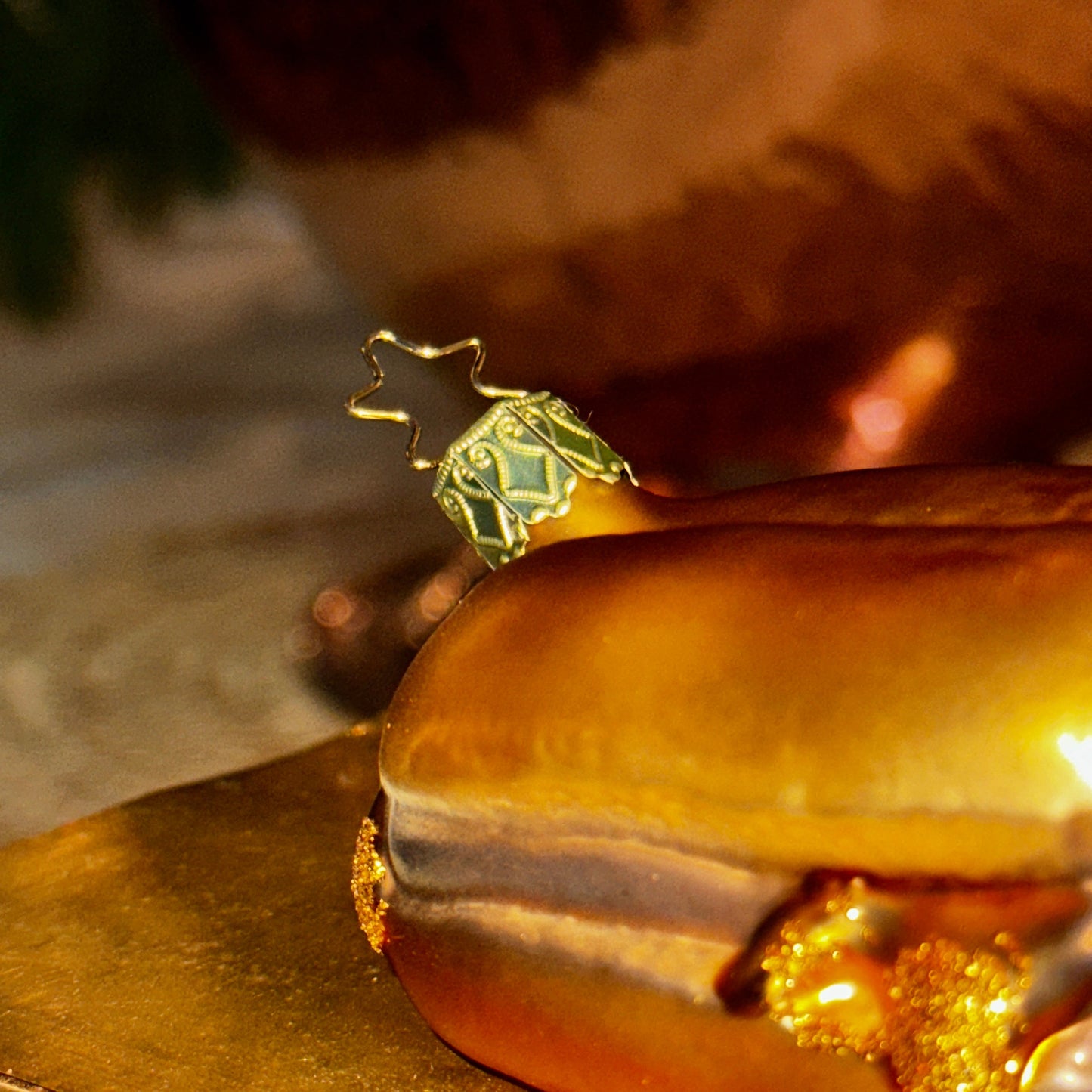 Nahaufnahme eines mundgeblasenen Christbaumschmucks in Form von "3 im Weggla". Der Fokus liegt auf der kunstvoll verzierten, goldfarbenen Aufhängung mit einem filigranen Stern-Detail. Das glänzende Brötchen mit den Bratwürsten schimmert in warmen Gold- und Brauntönen, akzentuiert durch feine Glitzerelemente. Im verschwommenen Hintergrund sorgen warme Lichtreflexe und goldene Dekorationen für eine festliche Atmosphäre.