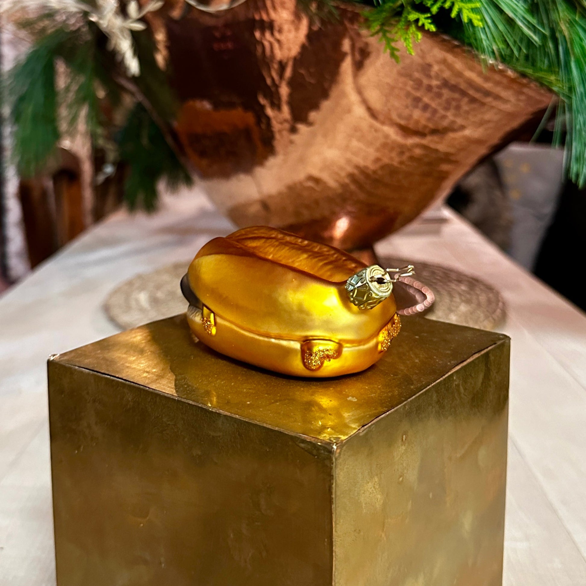 Ein mundgeblasener Christbaumschmuck in Form von "3 im Weggla" liegt auf einem goldenen Podest. Die Glasfigur zeigt ein glänzendes, goldfarbenes Brötchen mit fein modellierter Oberflächenstruktur. An den Seiten sind dezente Glitzerelemente zu sehen, die Senf oder Bratensaft symbolisieren. Die kunstvoll verzierte, goldene Aufhängung mit einem filigranen Stern-Detail ist sichtbar. Im Hintergrund sorgen grüne Tannenzweige und eine warme, festliche Beleuchtung für eine weihnachtliche Atmosphäre.