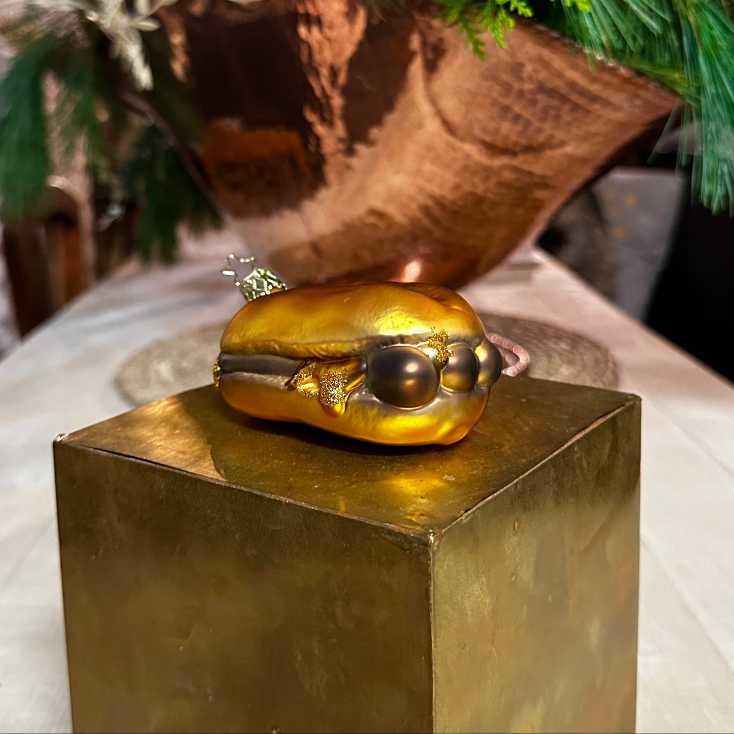 Ein mundgeblasener Christbaumschmuck in Form von "3 im Weggla" liegt auf einem goldenen Podest. Die glänzende Glasfigur stellt drei detailreiche Nürnberger Bratwürste in einem goldfarbenen Brötchen dar, verziert mit feinen Glitzerelementen, die Senf oder Bratensaft symbolisieren. Die filigrane, goldene Aufhängung mit Stern-Detail ist sichtbar. Im Hintergrund sorgen grüne Tannenzweige und eine warme Beleuchtung für eine festliche Weihnachtsstimmung.