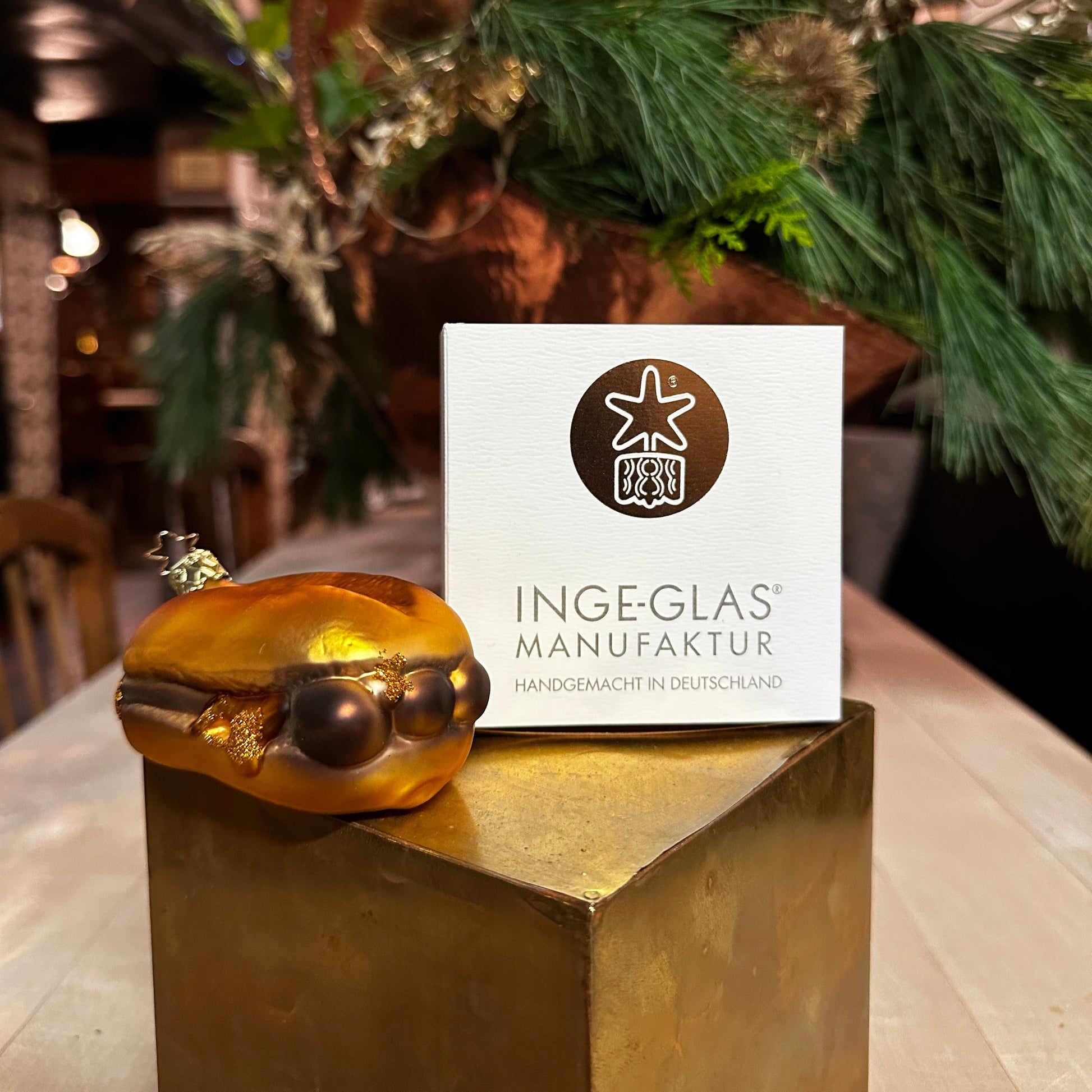 Ein mundgeblasener Christbaumschmuck in Form von "3 im Weggla" liegt auf einem goldenen Podest vor einer weißen Verpackung der Inge-Glas Manufaktur mit dem Schriftzug "Handgemacht in Deutschland". Die Glasfigur stellt drei Nürnberger Bratwürste in einem goldfarbenen Brötchen dar, mit liebevollen Details wie glänzenden Farbakzenten und glitzernden Highlights. Im Hintergrund sorgen grüne Tannenzweige und warme Lichtreflexe für eine festliche Weihnachtsatmosphäre.