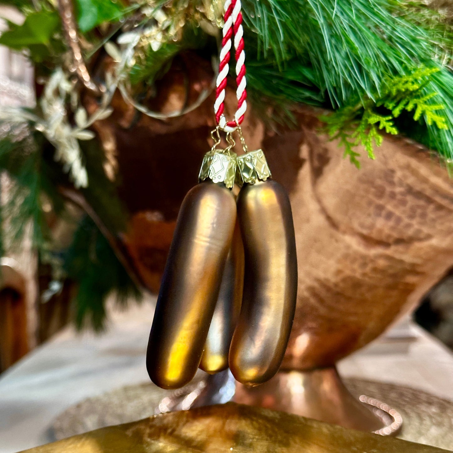 Drei glänzende, mundgeblasene Christbaumkugeln in Form von Nürnberger Bratwürsten hängen an einer rot-weiß gestreiften Schnur. Die handbemalten Würste haben eine realistische braune Färbung und sind mit filigranen, goldverzierten Aufhängern versehen. Im Hintergrund sorgen grüne Tannenzweige und eine kupferfarbene Dekoration für eine festliche, warme Atmosphäre. Die Bratwurst-Ornamente schweben leicht über einem goldenen Podest, das ihre edle Verarbeitung betont.