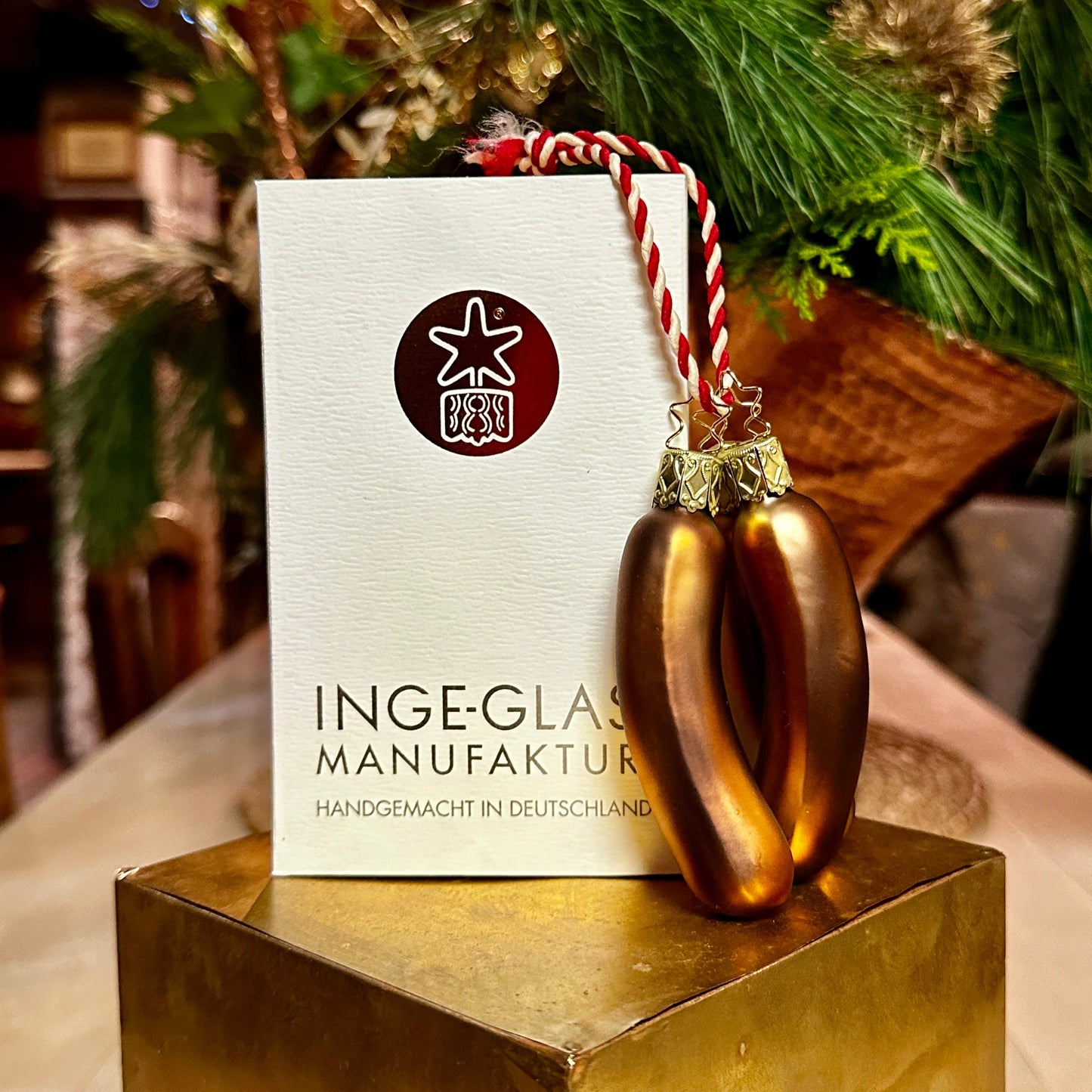 Ein handgefertigter Christbaumschmuck in Form von drei Nürnberger Bratwürsten hängt an einer rot-weiß gestreiften Schnur vor einer Verpackung der Inge-Glas Manufaktur mit dem Schriftzug "Handgemacht in Deutschland". Die glänzend braunen, mundgeblasenen Bratwürste haben detailreiche, goldverzierte Aufhänger und stehen auf einem goldenen Podest. Im Hintergrund sorgen grüne Tannenzweige für eine weihnachtliche Atmosphäre.