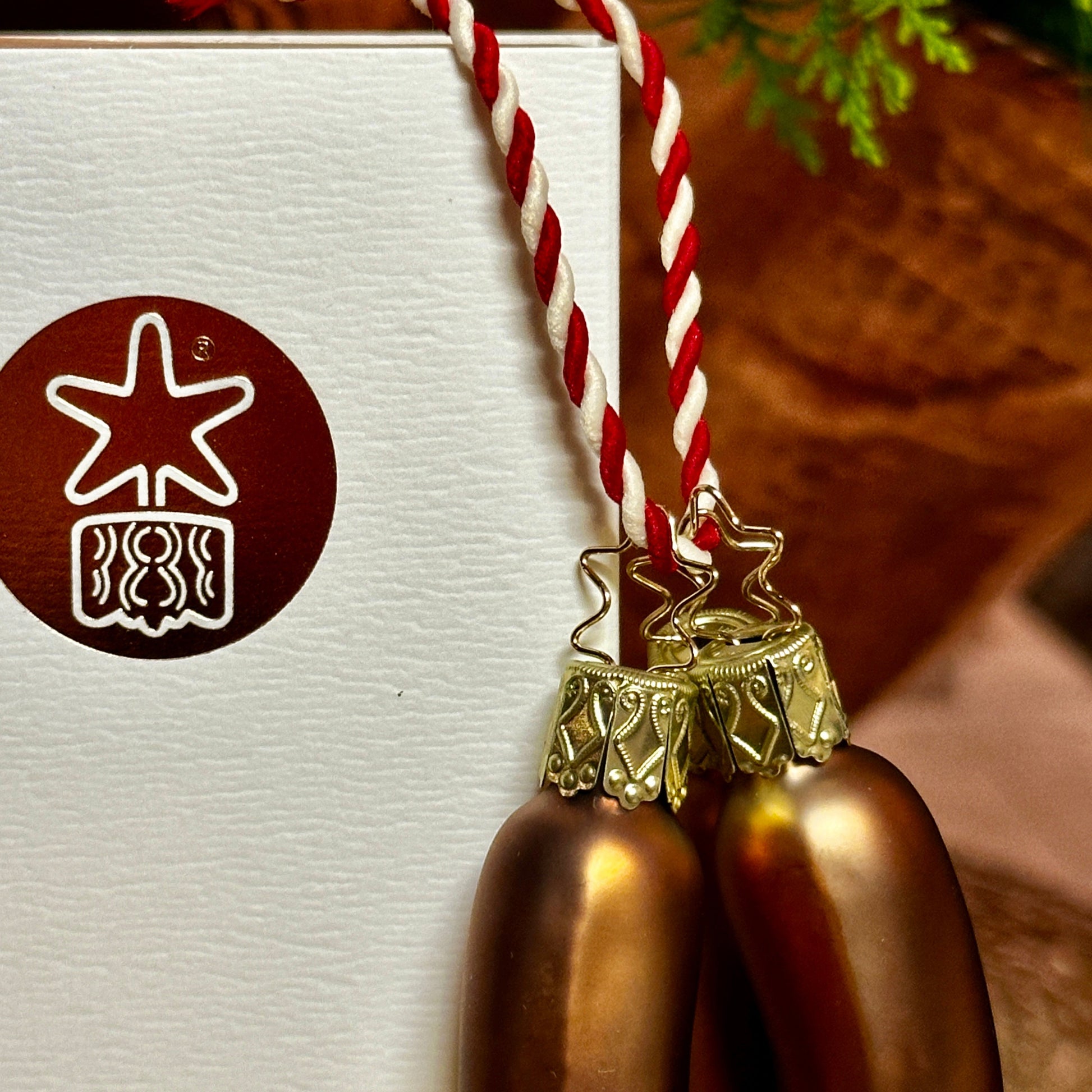 Nahaufnahme von drei mundgeblasenen Christbaumkugeln in Form von Nürnberger Bratwürsten, die an einer rot-weiß gedrehten Schnur hängen. Die handbemalten Bratwürste haben eine realistische, glänzende braune Färbung und sind mit kunstvoll verzierten, goldenen Aufhängern mit filigranen Stern-Details versehen. Im Hintergrund ist eine weiße Verpackung mit dem Logo der Inge-Glas Manufaktur zu sehen, die für hochwertige, handgefertigte Glasornamente steht.