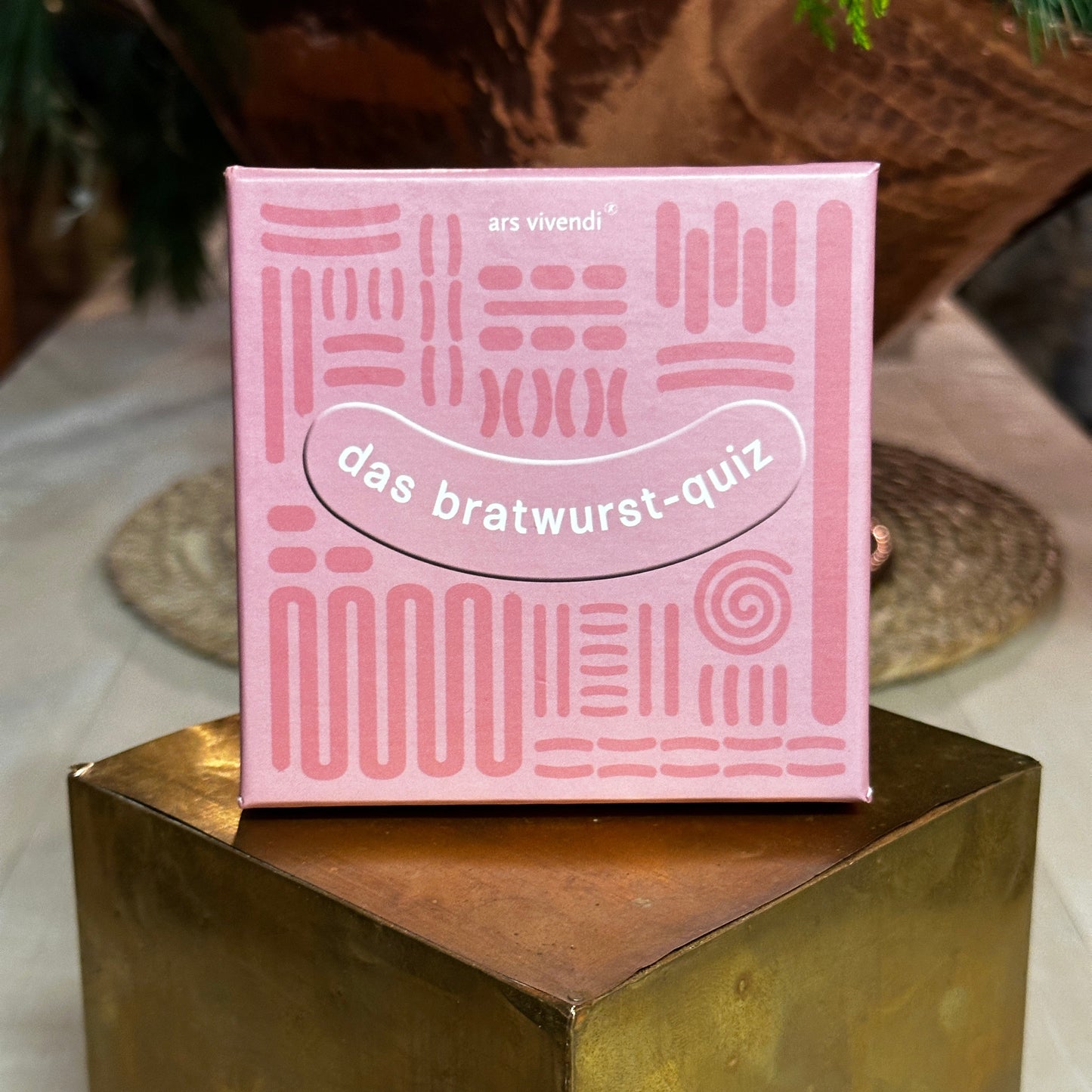 Ein rosa Karton mit der Aufschrift "das bratwurst-quiz" in einer Wurst-förmigen Grafik steht auf einem goldenen Podest. Das Design der Verpackung ist mit abstrakten Mustern verziert, die an Bratwurst und Grillmuster erinnern. Im Hintergrund ist eine gemütliche, leicht verschwommene Szenerie mit Naturmaterialien wie Holz und Geflecht zu sehen.