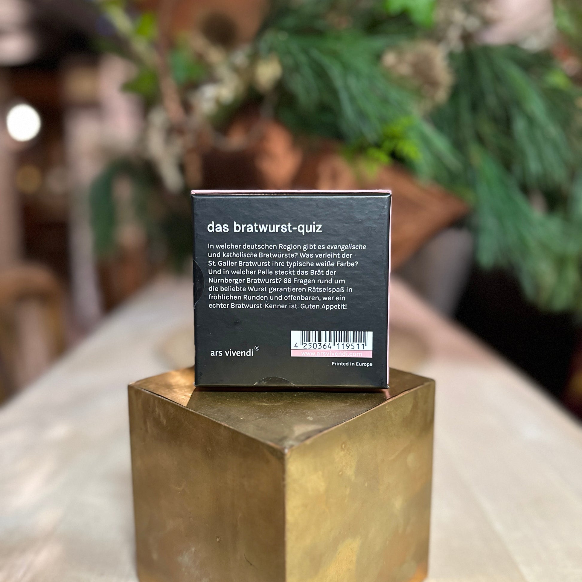 Die Rückseite der Verpackung des "Bratwurst-Quiz" ist zu sehen. Der schwarze Karton enthält eine kurze Beschreibung des Spiels mit Fragen rund um die Bratwurst, z. B. zu regionalen Unterschieden und speziellen Zutaten. Unten befindet sich ein Barcode sowie das Logo des Verlags ars vivendi. Die Verpackung steht auf einem goldenen Podest, während der Hintergrund mit grünen Pflanzen und einer gemütlichen, leicht unscharfen Atmosphäre gestaltet ist.