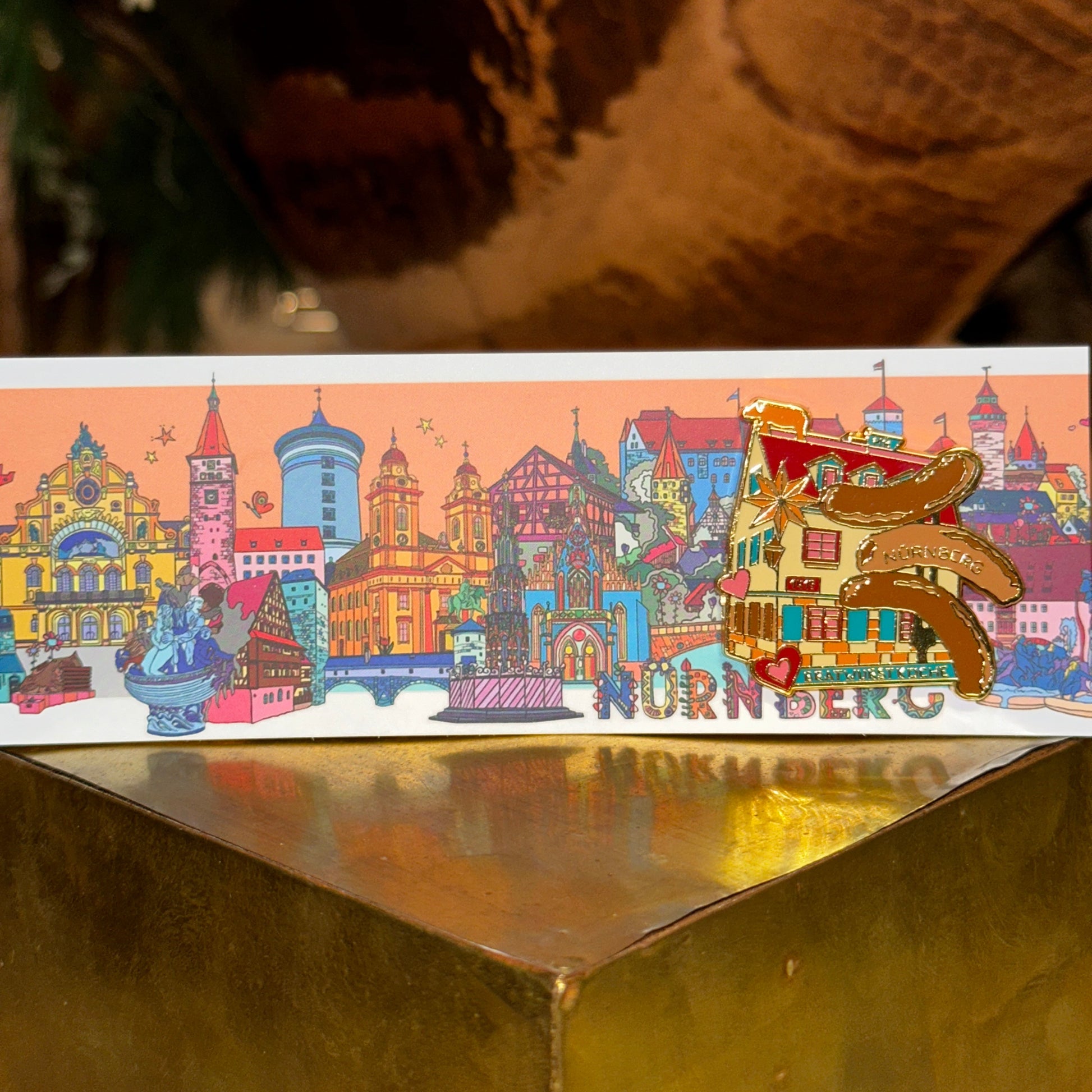Ein kunstvoll gestalteter Ansteck-Pin mit der Aufschrift "Bratwurstküche Nürnberg" ist auf einer bunten Postkarte befestigt. Der Pin zeigt eine stilisierte Darstellung eines historischen Gebäudes mit roten Fensterläden, goldenen Akzenten und mehreren gegrillten Bratwürsten mit der Aufschrift "Nürnberg". Liebevolle Details wie Herzen und Sterne schmücken das Motiv. Der Hintergrund zeigt eine farbenfrohe Illustration von Nürnberger Wahrzeichen, darunter Türme, Fachwerkhäuser und historische Gebäude. Der Pin l