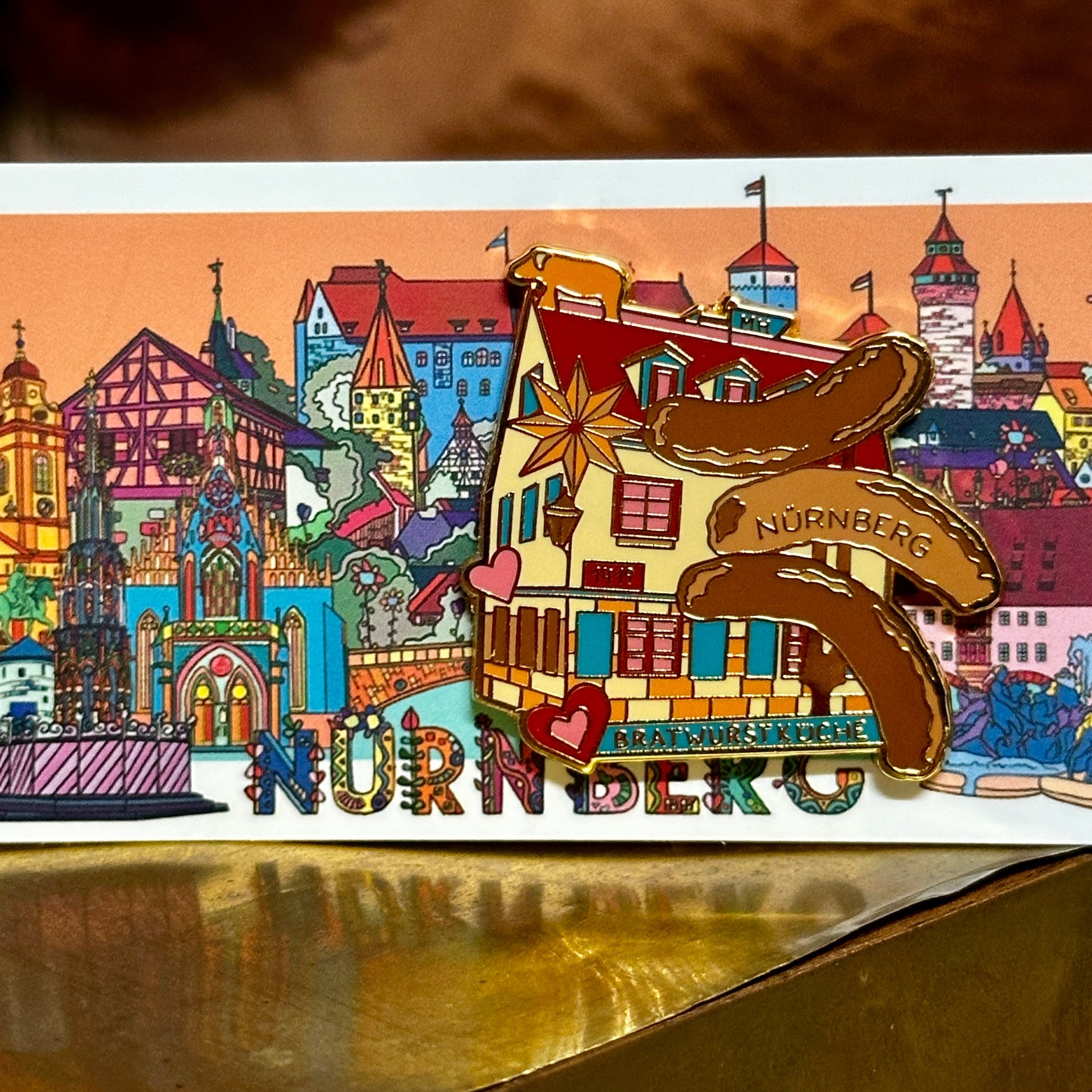 Ein farbenfroher Ansteck-Pin mit der Aufschrift "Bratwurstküche Nürnberg" ist auf einer Postkarte mit einer detailreichen Illustration von Nürnberger Wahrzeichen befestigt. Der Pin zeigt ein traditionelles Fachwerkhaus mit roten Fensterläden, goldenen Akzenten und drei geschwungenen Bratwürsten, von denen eine die Aufschrift "Nürnberg" trägt. Ein kleines Herz sowie ein goldener Stern verzieren das Design. Die Postkarte im Hintergrund präsentiert eine bunte Skyline mit bekannten Gebäuden.