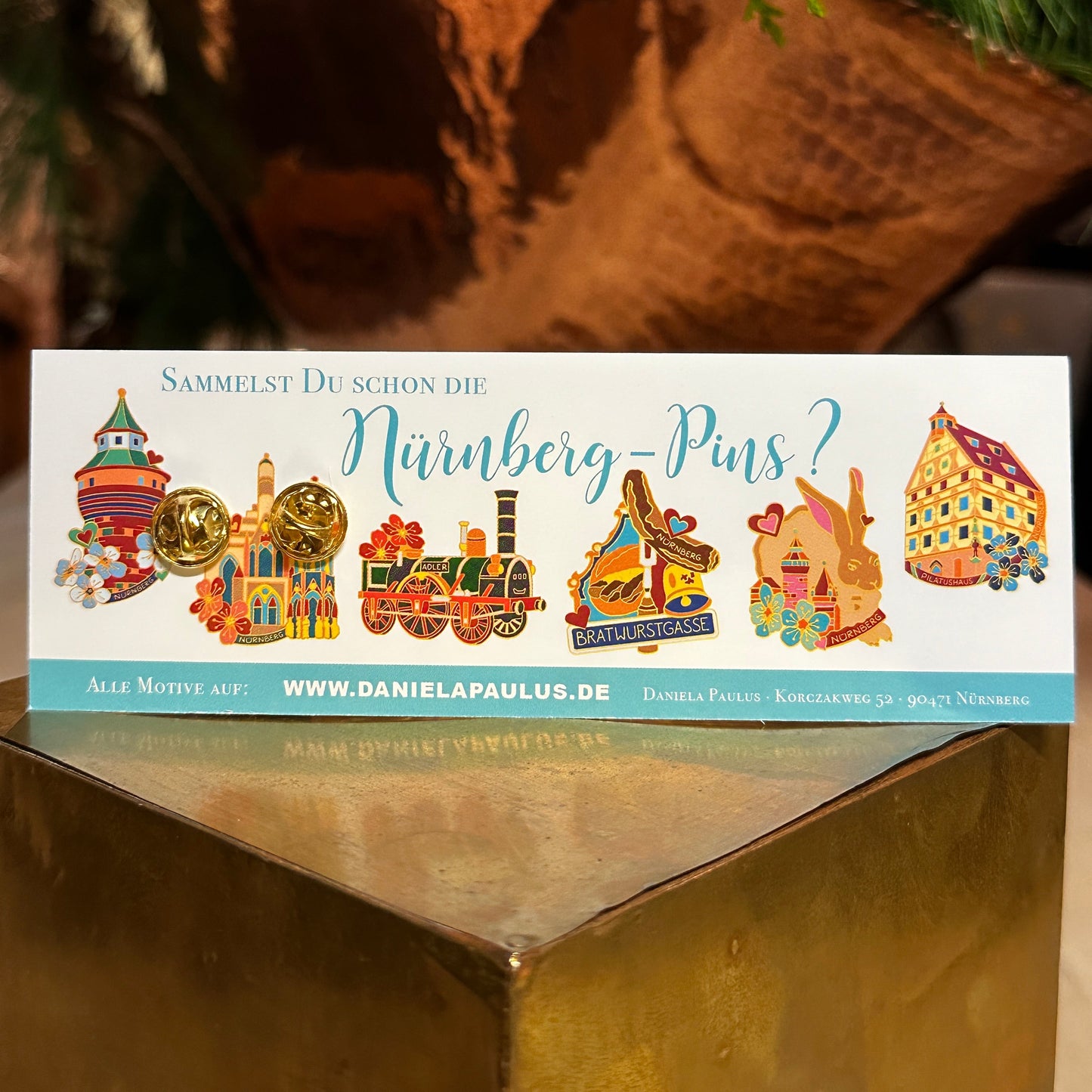 Eine Sammelkarte mit der Aufschrift "Sammelst Du schon die Nürnberg-Pins?" präsentiert verschiedene kunstvolle Ansteck-Pins mit Motiven aus Nürnberg. Die Karte zeigt liebevoll illustrierte Designs, darunter den runden Turm der Stadt, eine gotische Kirche, eine historische Dampflok, den Bratwurstgassen-Pin, einen Hasen mit Herzdetails und ein traditionelles Fachwerkhaus. Zwei goldene Pin-Verschlüsse sind auf der linken Seite befestigt. Die Karte verweist auf die Webseite www.danielapaulus.de