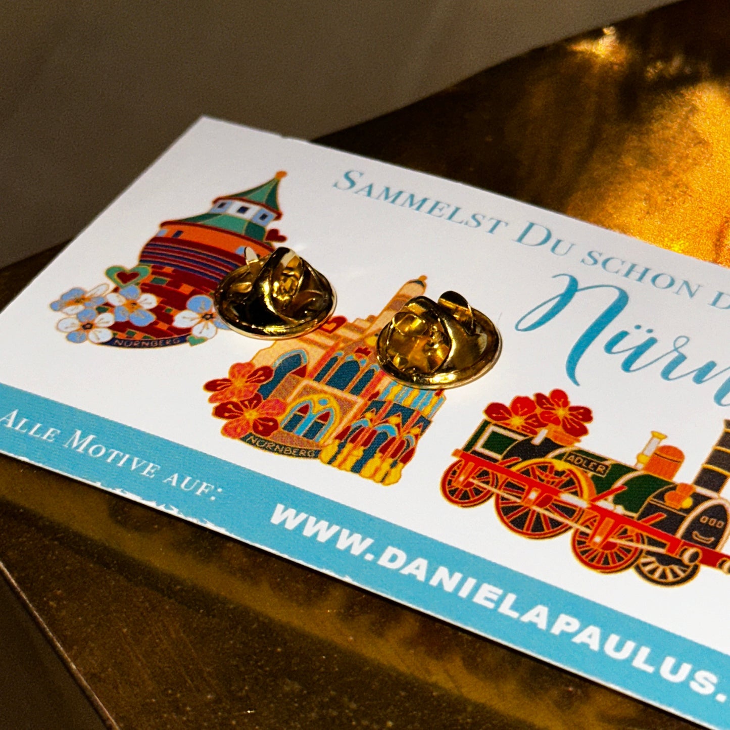 Die Rückseite eines goldfarbenen Ansteck-Pins ist auf einer illustrierten Sammelkarte befestigt. Die Karte zeigt bunte Motive von Nürnberger Wahrzeichen, darunter einen Turm, eine historische Kirche und eine Dampflok mit floralen Verzierungen. Der Text auf der Karte beginnt mit "Sammelst du schon..." und verweist auf die Webseite www.danielapaulus.com, die weitere Motive der Pin-Kollektion präsentiert. Das Bild ist auf einem goldenen Untergrund aufgenommen, der die edle Optik des Pins unterstreicht.