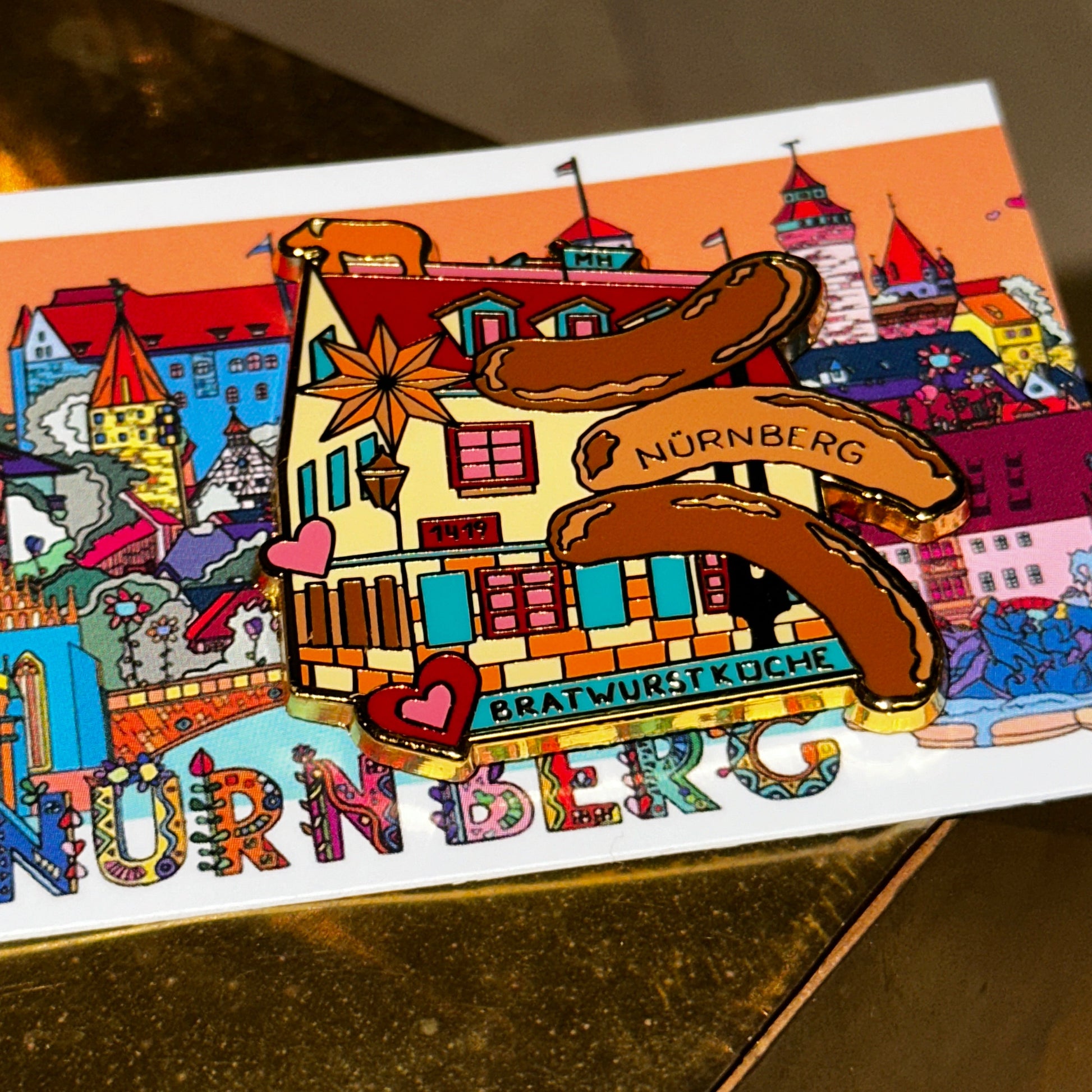 Ein kunstvoll gestalteter Ansteck-Pin mit der Aufschrift "Bratwurstküche Nürnberg" ist auf einer bunten Postkarte befestigt. Der Pin zeigt eine stilisierte Darstellung eines historischen Gebäudes mit roten Fensterläden, goldenen Akzenten und mehreren gegrillten Bratwürsten mit der Aufschrift "Nürnberg". Liebevolle Details wie Herzen und Sterne schmücken das Motiv. Der Hintergrund zeigt eine farbenfrohe Illustration von Nürnberger Wahrzeichen, darunter Türme, Fachwerkhäuser und historische Gebäude. Der Pin