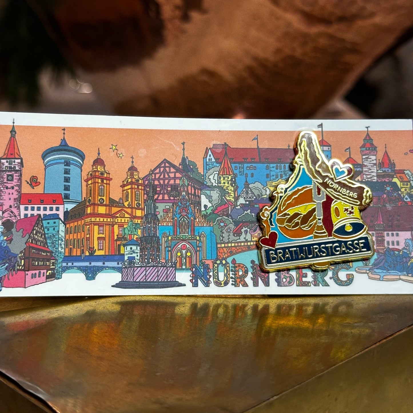 Ein kunstvoll gestalteter Ansteck-Pin mit der Aufschrift "Bratwurstgasse Nürnberg" ist auf einer Postkarte mit einer bunten Illustration der Stadt Nürnberg befestigt. Der Pin zeigt ein traditionelles Wirtshaus mit Bratwürsten auf einem Teller, dekoriert mit goldenen Akzenten und liebevollen Details. Der Hintergrund besteht aus warmen Farben, während das Schmuckstück auf einem goldfarbenen Podest präsentiert wird.