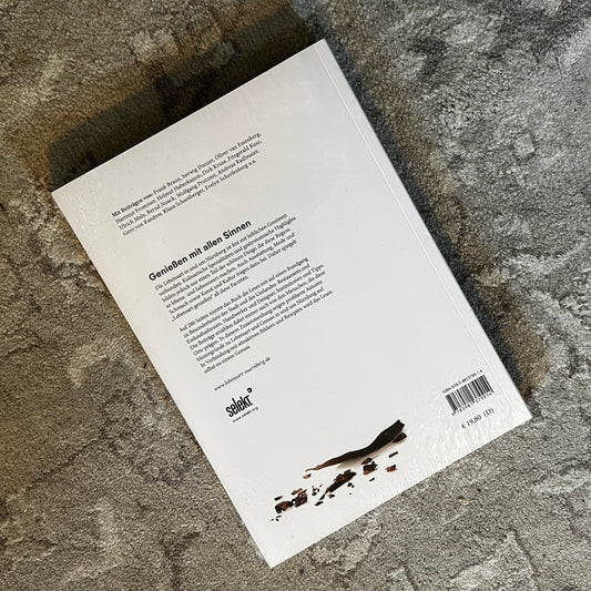 Rückseite eines Buches mit weißem Cover, das auf einem Teppich liegt. Der Titel "Genießen mit allen Sinnen" ist in schwarzer Schrift abgedruckt, begleitet von einer ausführlichen Beschreibung des Inhalts. Im unteren Bereich befindet sich das Logo des Verlags "selekkt" sowie der Preis von 19,80 €.