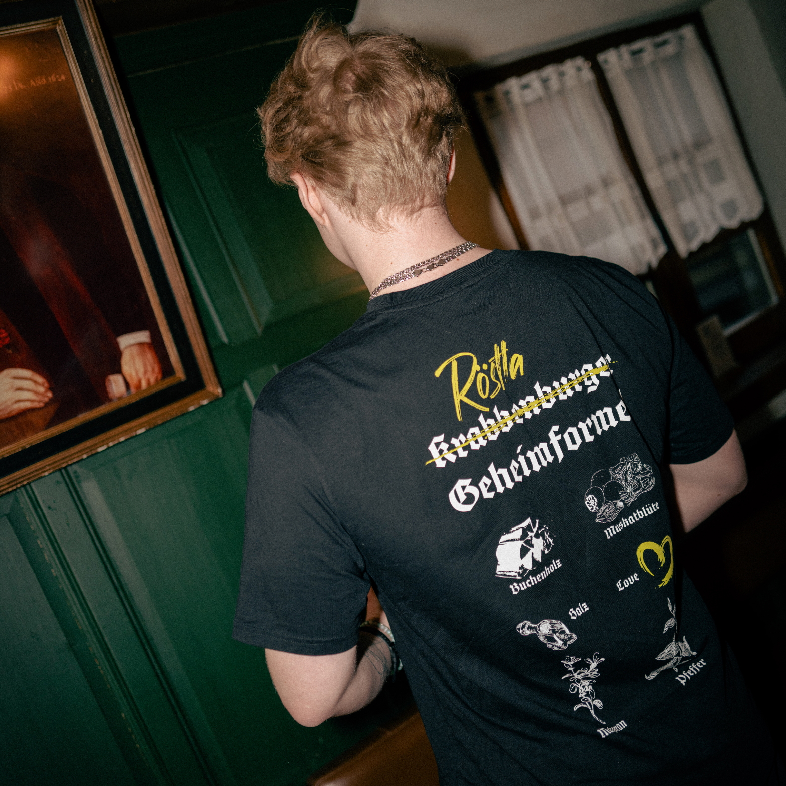 Eine Person mit kurzen, welligen, blonden Haaren steht mit dem Rücken zur Kamera in einer rustikalen Gaststube. Sie trägt ein schwarzes „Röstla Geheimformel“ T-Shirt, auf dessen Rückseite in grüner und weißer Schrift Zutaten wie Buchholz, Muskatblüte, Salz, Pfeffer und „Love“ abgebildet sind. Der Begriff „Krabbenburger“ ist durchgestrichen, wodurch der fränkische Humor des Designs betont wird. Im Hintergrund sind grüne Holzwände, eine Sitzbank aus braunem Leder, ein altes Gemälde und eine Regalreihe.