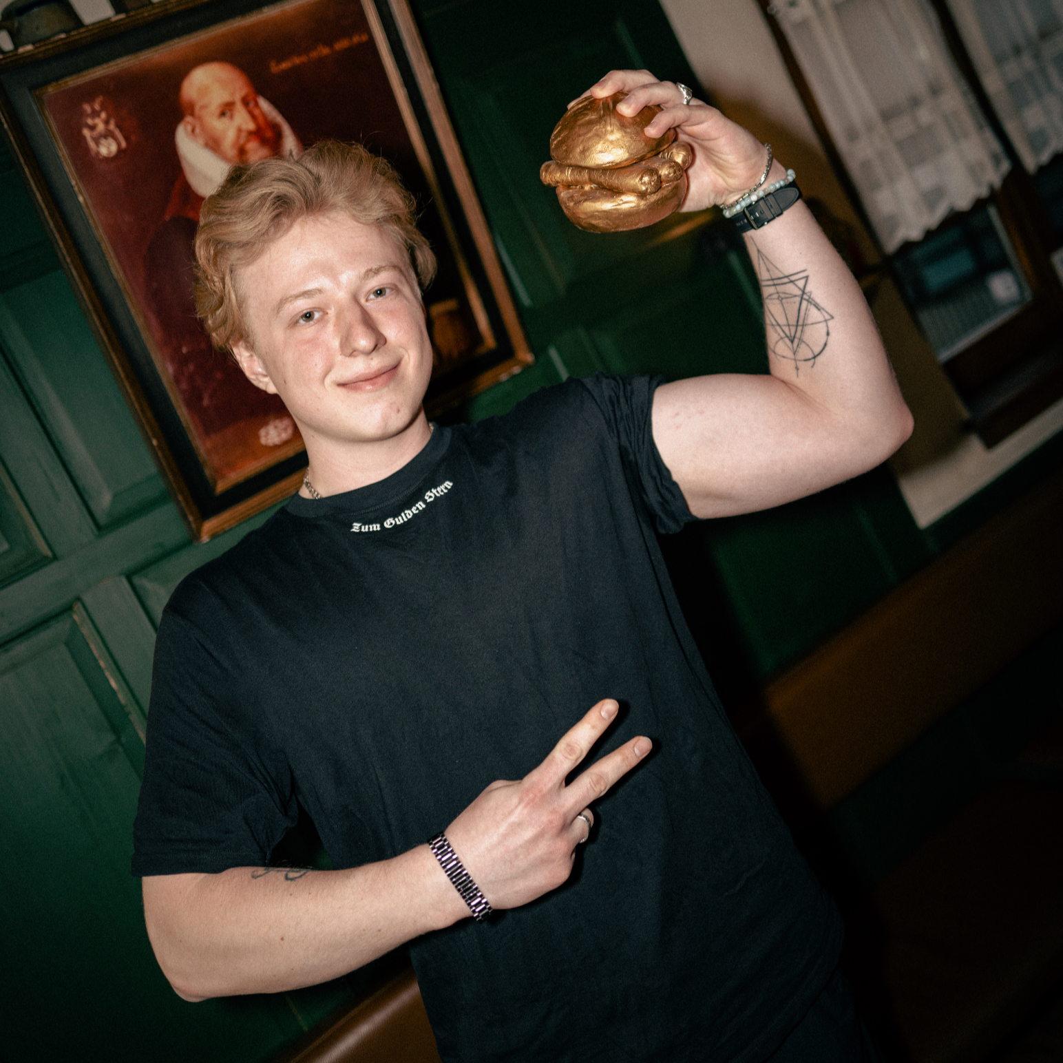 Ein junger Mann mit welligem, blondem Haar und einem leichten Lächeln trägt ein schwarzes T-Shirt mit der weißen Aufschrift „Zum Gulden Stern“ am Kragen. Er hält eine goldene Bratwurst im Brötchen in die Höhe und zeigt mit der anderen Hand ein Victory-Zeichen. Sein Arm ist mit einem geometrischen Tattoo verziert, und er trägt mehrere Armbänder. Im Hintergrund sind grüne Holzwände, eine braune Lederbank, ein altes Gemälde und eine Regalreihe mit traditionellen Keramikkrügen zu sehen.