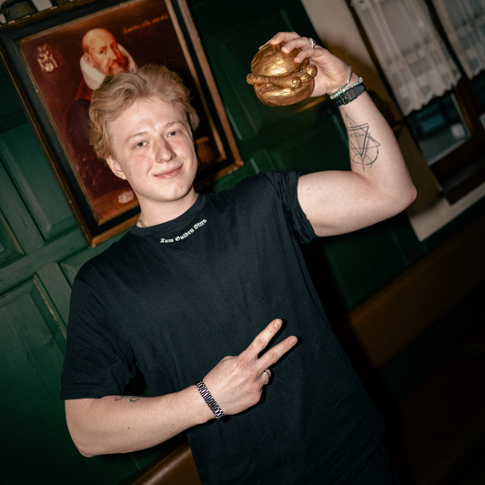 Ein junger Mann mit welligem, blondem Haar und einem leichten Lächeln trägt ein schwarzes T-Shirt mit der weißen Aufschrift „Zum Gulden Stern“ am Kragen. Er hält eine goldene Bratwurst im Brötchen in die Höhe und zeigt mit der anderen Hand ein Victory-Zeichen. Sein Arm ist mit einem geometrischen Tattoo verziert, und er trägt mehrere Armbänder. Im Hintergrund sind grüne Holzwände, eine braune Lederbank, ein altes Gemälde und eine Regalreihe mit traditionellen Keramikkrügen zu sehen.