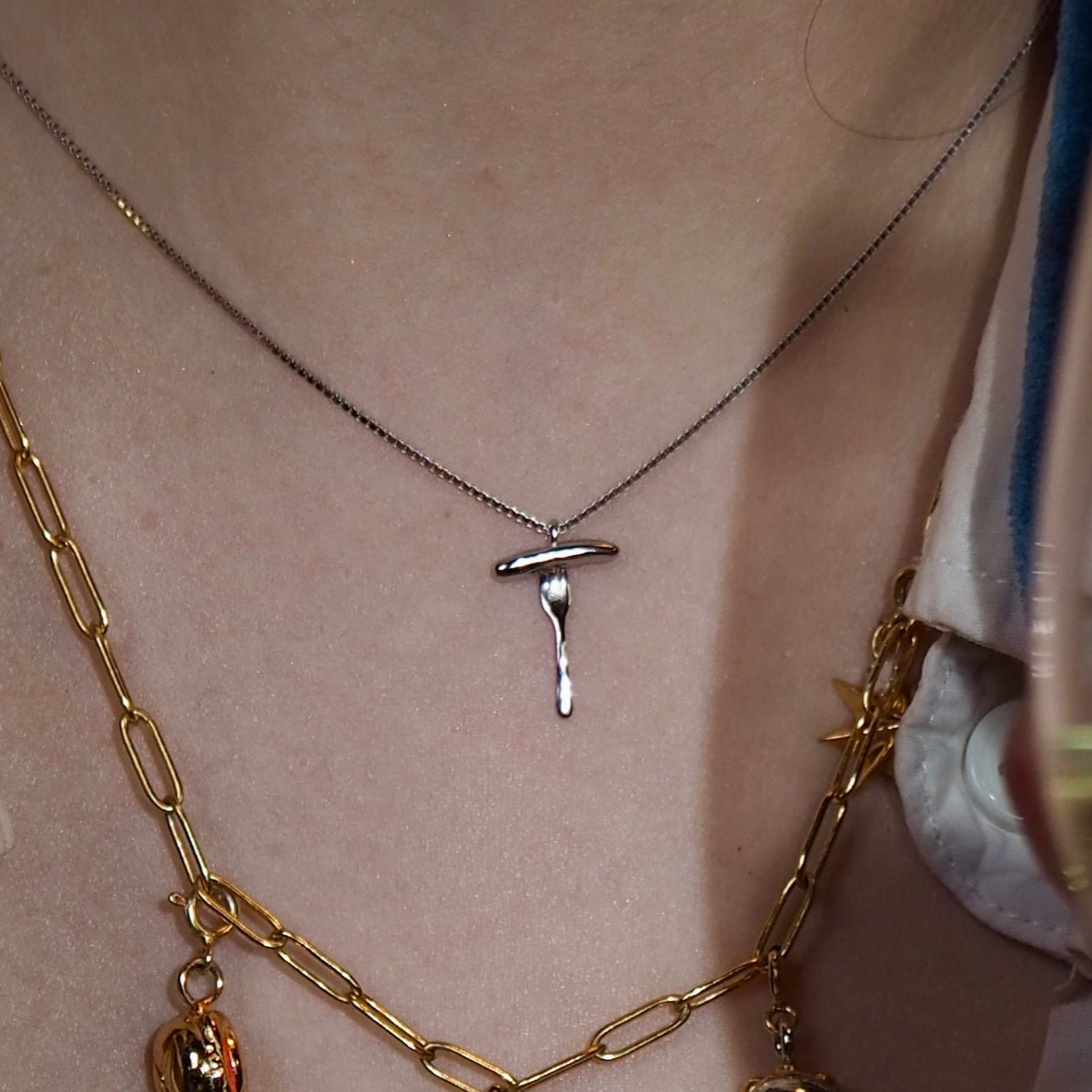 Nahaufnahme einer Halskette mit einem kleinen Silberanhänger in Form einer Bratwurst auf einer Gabel. Die Kette liegt auf der Haut einer Person, die zudem eine goldene Kette mit verschiedenen Anhängern trägt. Das Bild hat einen warmen, edlen Look, während im Vordergrund ein Glas mit einem Getränk zu sehen ist.