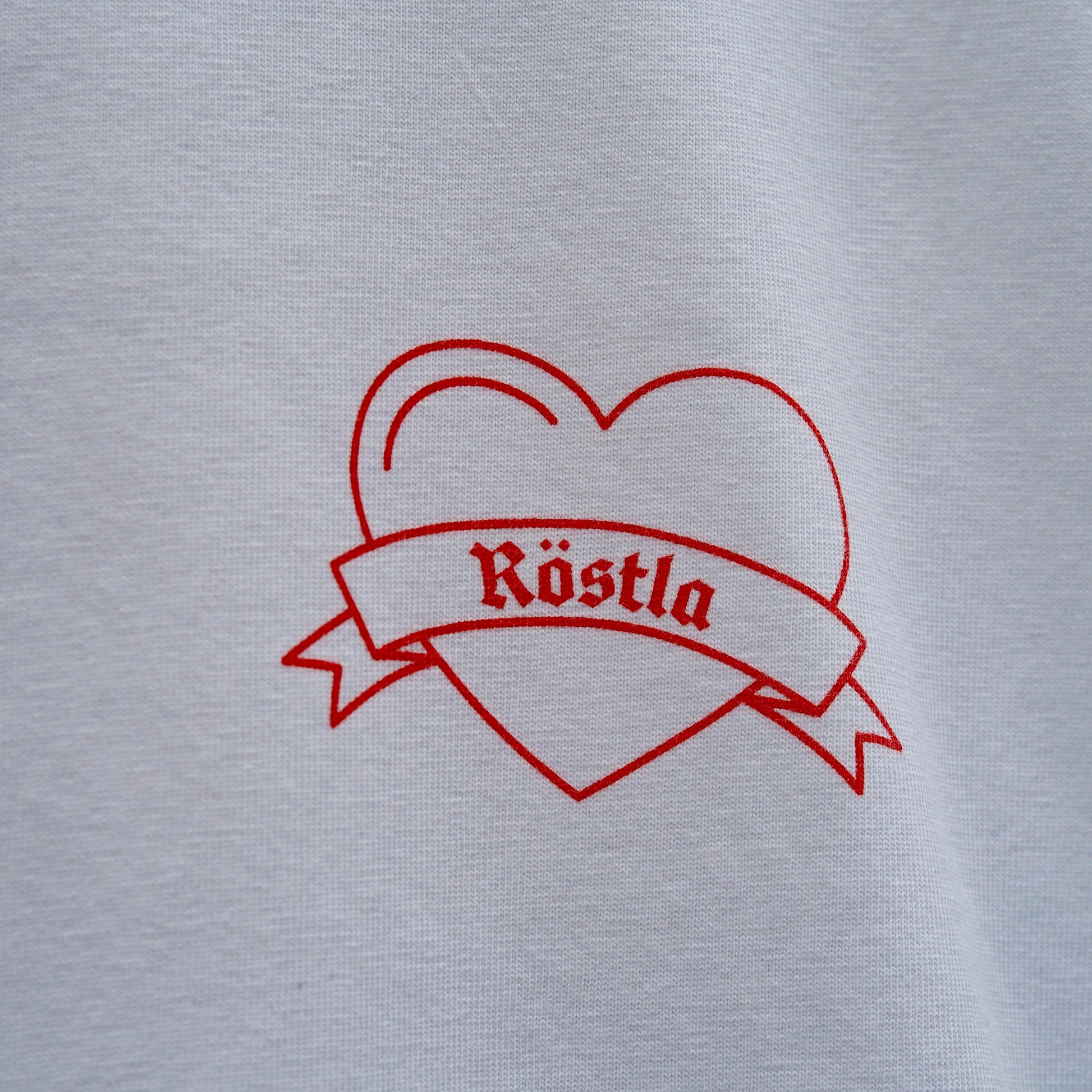 Nahaufnahme eines weißen T-Shirts mit einem roten, linearen Print. Das Motiv zeigt ein Herz mit einer geschwungenen Schleife, auf der in gotischer Schrift das Wort „Röstla“ steht. Das Design wirkt minimalistisch und gleichzeitig traditionell, inspiriert von klassischen Tattoo-Motiven.
