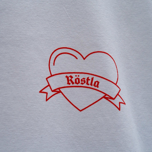 Nahaufnahme eines weißen T-Shirts mit einem roten, linearen Print. Das Motiv zeigt ein Herz mit einer geschwungenen Schleife, auf der in gotischer Schrift das Wort „Röstla“ steht. Das Design wirkt minimalistisch und gleichzeitig traditionell, inspiriert von klassischen Tattoo-Motiven.