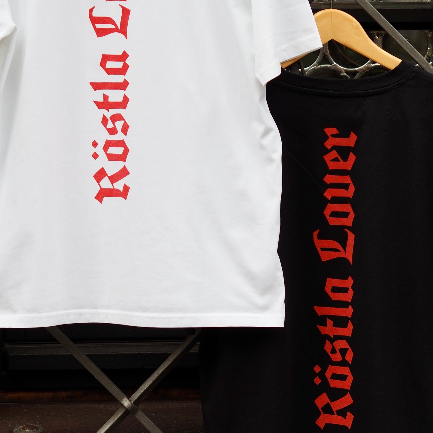 Zwei T-Shirts mit dem Aufdruck „Röstla Lover“ in roter gotischer Schrift. Ein weißes T-Shirt hängt vorne, während ein schwarzes T-Shirt dahinter auf einem Kleiderbügel hängt. Die Schrift verläuft vertikal über den Rücken der Shirts und verleiht ihnen einen markanten, modernen Look mit traditionellem Flair.