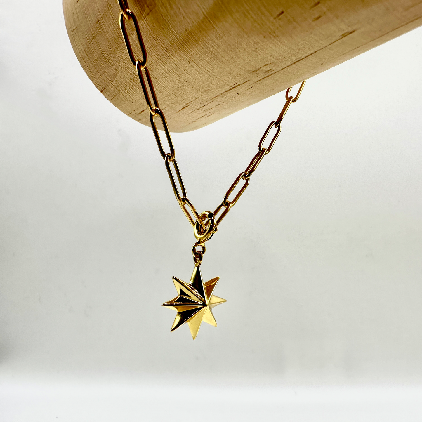 Vergoldetes Sammelarmband mit grober Gliederkette und einem filigranen, achtzackigen Stern-Anhänger. Das Armband ist dekorativ über eine hölzerne Stange drapiert, wodurch die hochwertige Verarbeitung und das edle Design besonders zur Geltung kommen.