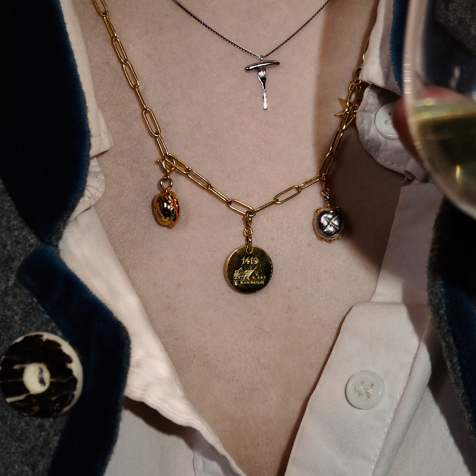 Eine stilvolle goldene Gliederkette mit mehreren individuellen Anhängern wird an einem Hals getragen. Die Kette besteht aus rechteckigen Gliedern und ist mit verschiedenen Schmuckstücken verziert, darunter ein runder 1419-Medaillon-Anhänger, ein silber-goldener „Drei im Weggla“-Anhänger, ein kleiner goldener Anhänger in Form eines Brötchens mit Bratwurst und ein Stern-Anhänger. Darüber liegt eine schlichte, silberne Kette mit einem Bratwurst-auf-Gabel-Anhänger. Die Person trägt eine weiße Bluse.
