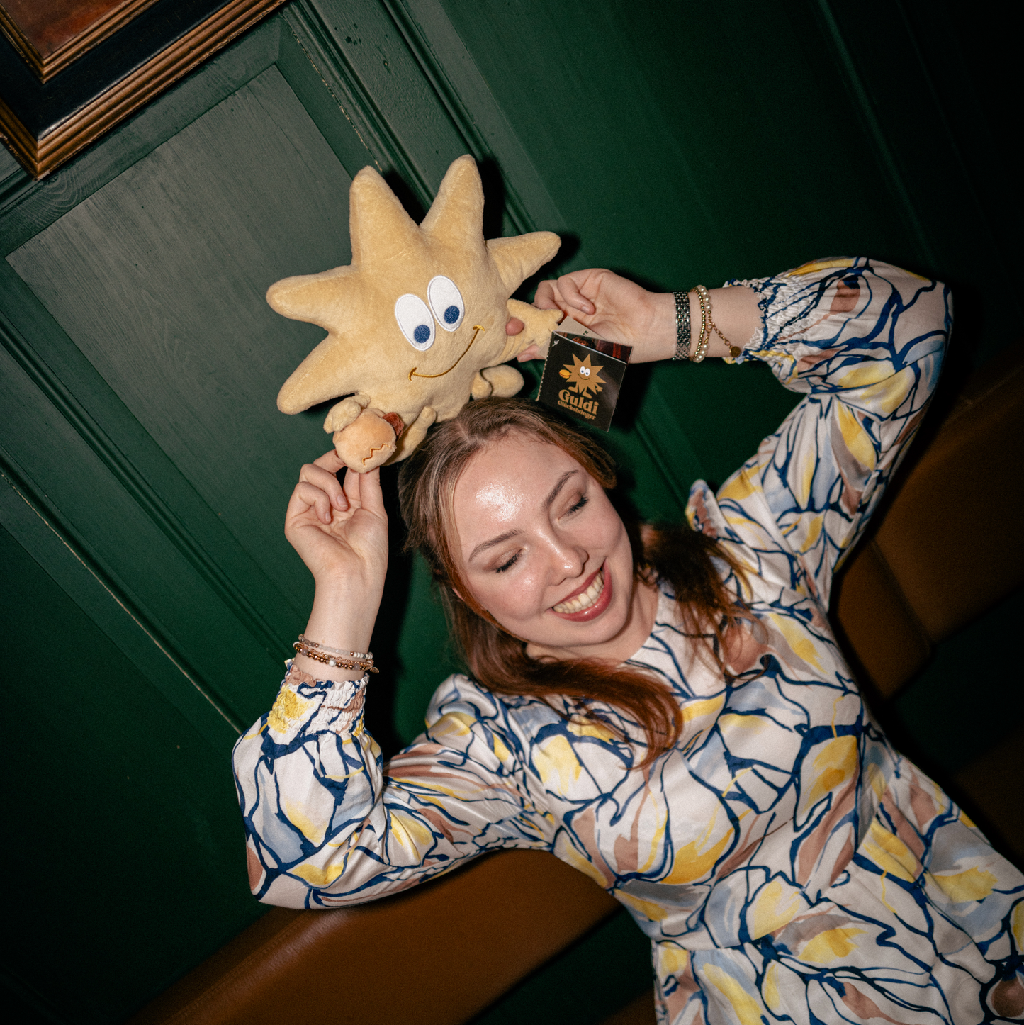 Eine fröhliche Frau mit welligen, rötlich-blonden Haaren hält das Guldi Kuscheltier, einen lächelnden Stern mit 3 im Weggla in der Hand, über ihren Kopf. Sie trägt ein farbenfrohes, gemustertes Kleid mit langen Ärmeln und Schmuck an ihren Handgelenken. Die Szene spielt sich in einer gemütlichen, rustikalen Umgebung mit grüner Holzwand und brauner Sitzbank ab. Guldi’s Etikett ist sichtbar und zeigt seinen Namen als Glücksbringer.