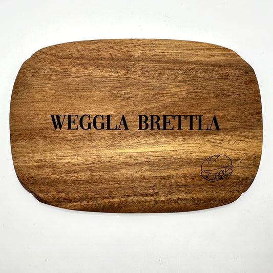 Ein hochwertiges Schneidebrett aus Holz mit der Gravur „Weggla Brettla“ in schwarzer Schrift. In der unteren rechten Ecke ist eine feine Gravur eines Brötchens mit drei Bratwürsten zu sehen. Das Brett hat eine abgerundete Form und eine warme Holzmaserung, die dem Design eine rustikale und authentische Note verleiht. Perfekt als Servierbrett oder stilvolles Küchenaccessoire für Liebhaber fränkischer Spezialitäten.