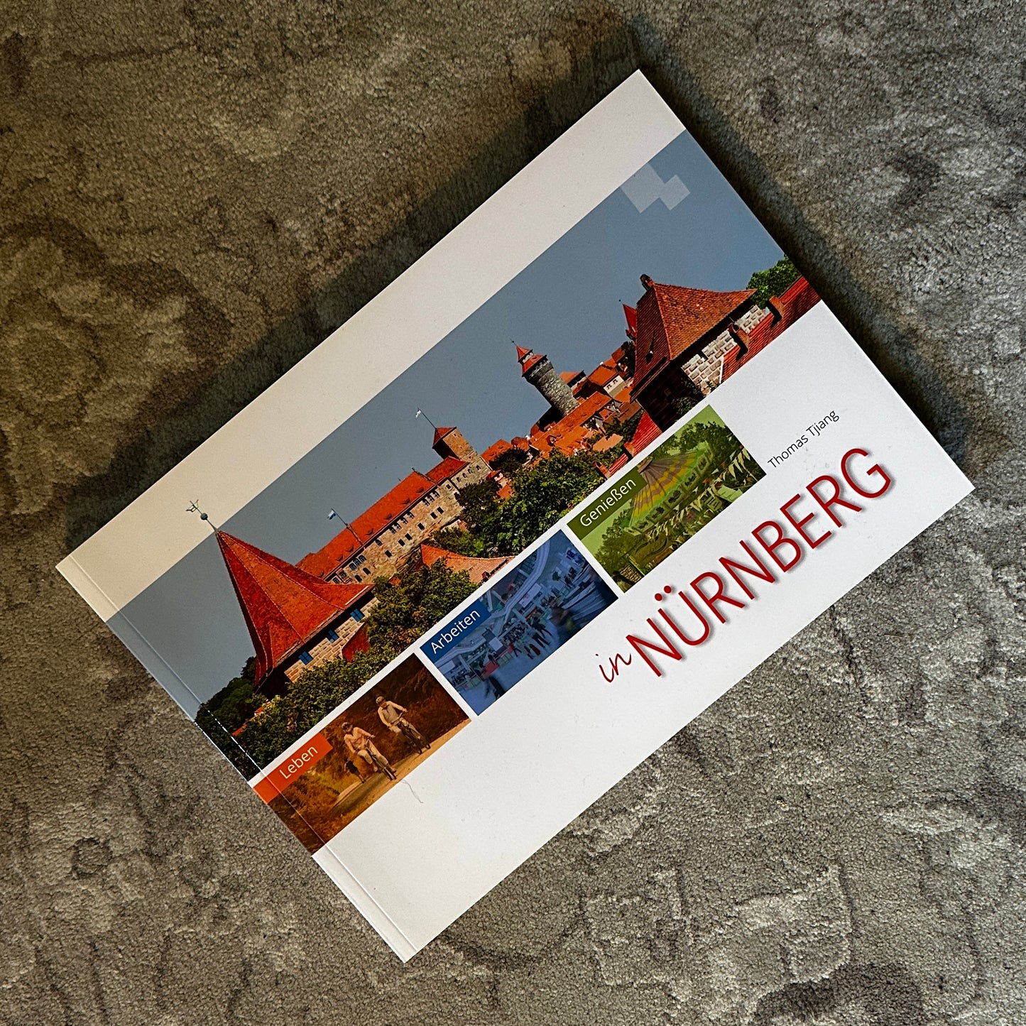 Das Buch „In Nürnberg“ von Thomas Tjiang liegt auf einem Teppich. Das Cover zeigt eine Collage mit Bildern der Nürnberger Burg, dem Stadtleben, Arbeitsumgebungen und kulinarischen Erlebnissen. Der Titel ist in markantem Rot gehalten, und das Buch bietet eine umfassende Darstellung des Lebens in der Stadt.