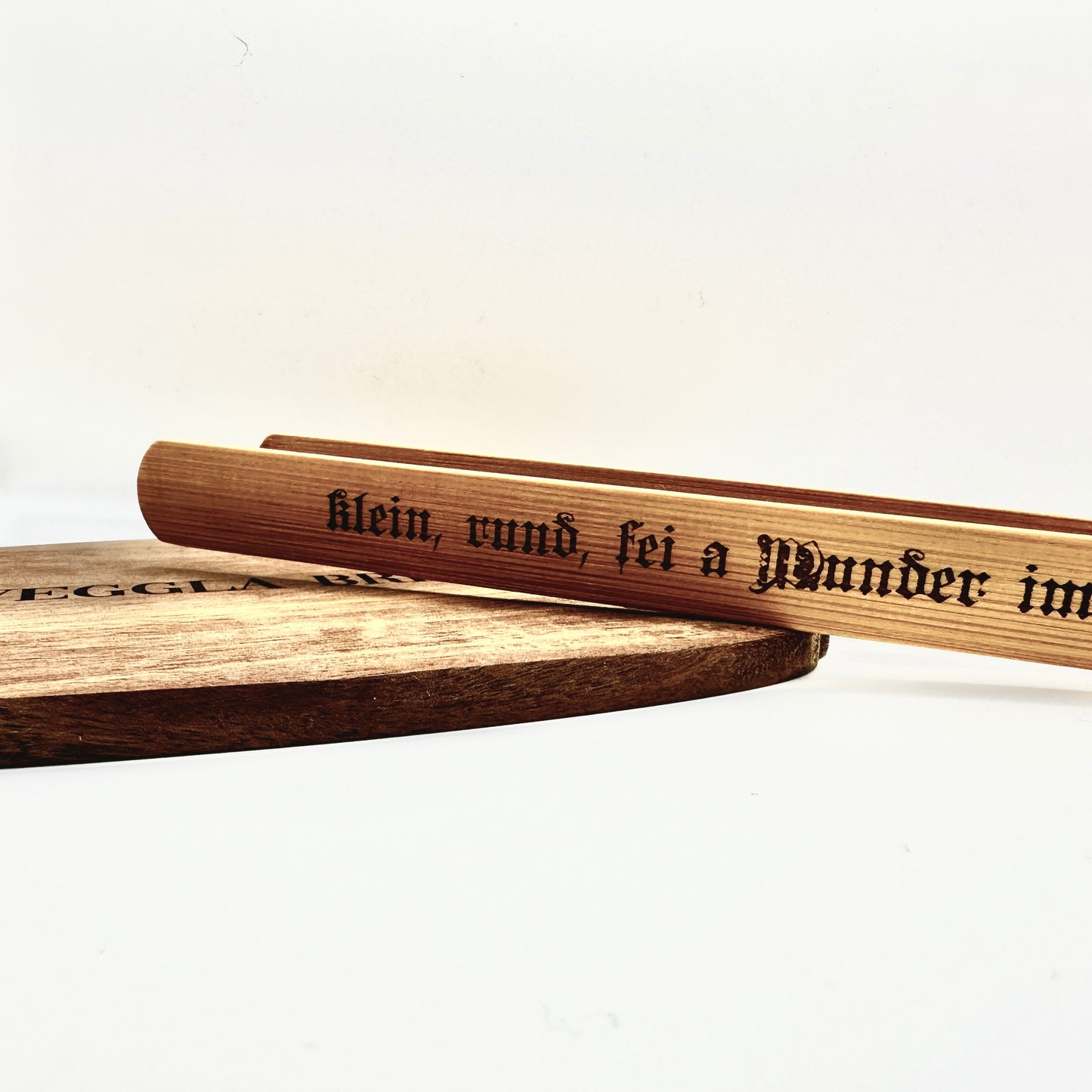Die Holzzange mit Gravur liegt auf einem rustikalen Holzbrett, was ihre traditionelle und handwerkliche Verarbeitung unterstreicht. Ein stilvolles Küchenaccessoire mit regionalem Charme.