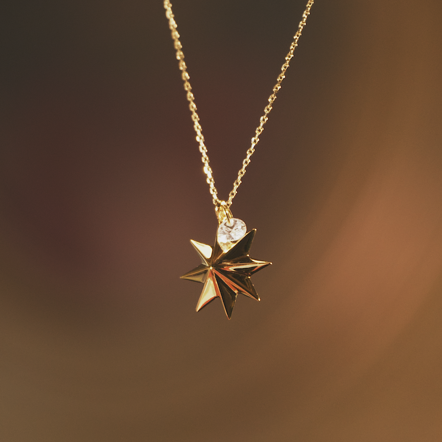 Nahaufnahme einer filigranen goldenen Halskette mit einem facettierten Stern-Anhänger in edler 3 Micron Vergoldung auf Sterling-Silber, ergänzt durch einen funkelnden Zirkonia-Stein. Der warme, unscharfe Hintergrund betont das elegante Design.