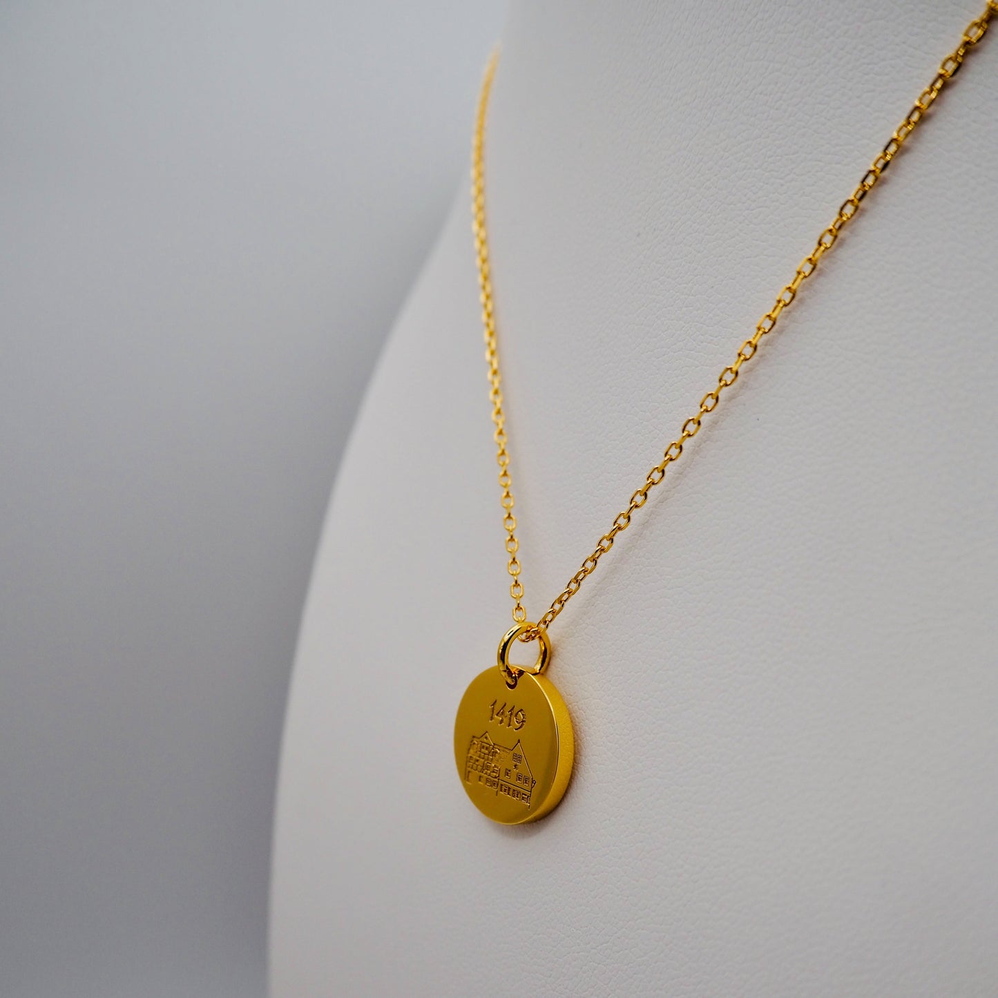 Nahaufnahme einer eleganten Goldkette mit rundem, gravierten Anhänger. Die Gravur zeigt das historische Gebäude der Bratwurstküche „Zum Gulden Stern“ mit der Jahreszahl „1419“. Das Schmuckstück hängt an einer feinen, vergoldeten Kette und wird stilvoll auf einem weißen Schmuckdisplay präsentiert.