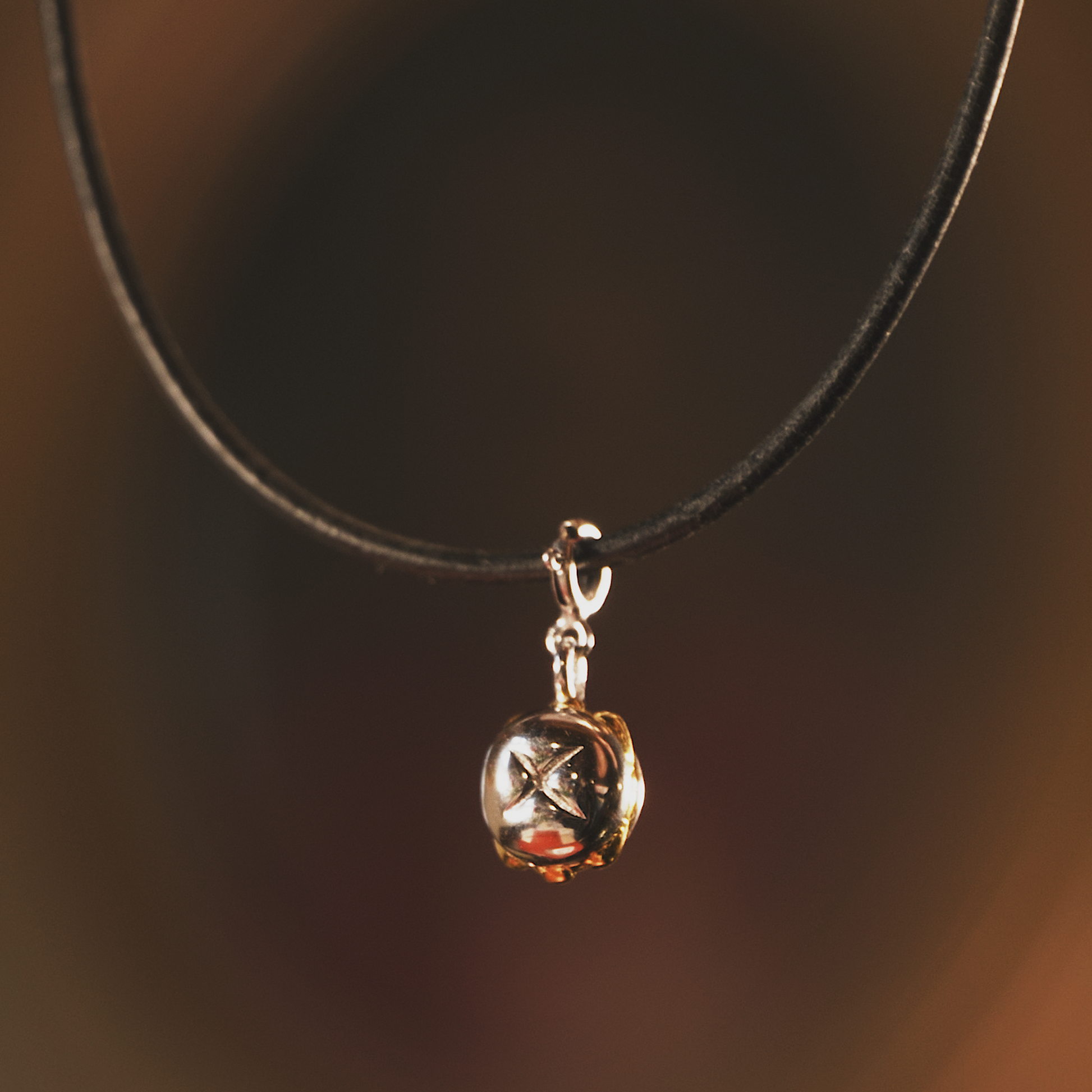 Nahaufnahme einer schwarzen Lederkette mit einem hochwertigen Anhänger in Form einer kleinen, glänzenden Glocke. Der Anhänger besteht aus echtem Sterling-Silber mit 3 Micron Vergoldung und zeigt feine Details. Der Hintergrund ist warm und unscharf, wodurch der Anhänger elegant in Szene gesetzt wird.