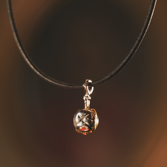 Nahaufnahme einer schwarzen Lederkette mit einem hochwertigen Anhänger in Form einer kleinen, glänzenden Glocke. Der Anhänger besteht aus echtem Sterling-Silber mit 3 Micron Vergoldung und zeigt feine Details. Der Hintergrund ist warm und unscharf, wodurch der Anhänger elegant in Szene gesetzt wird.