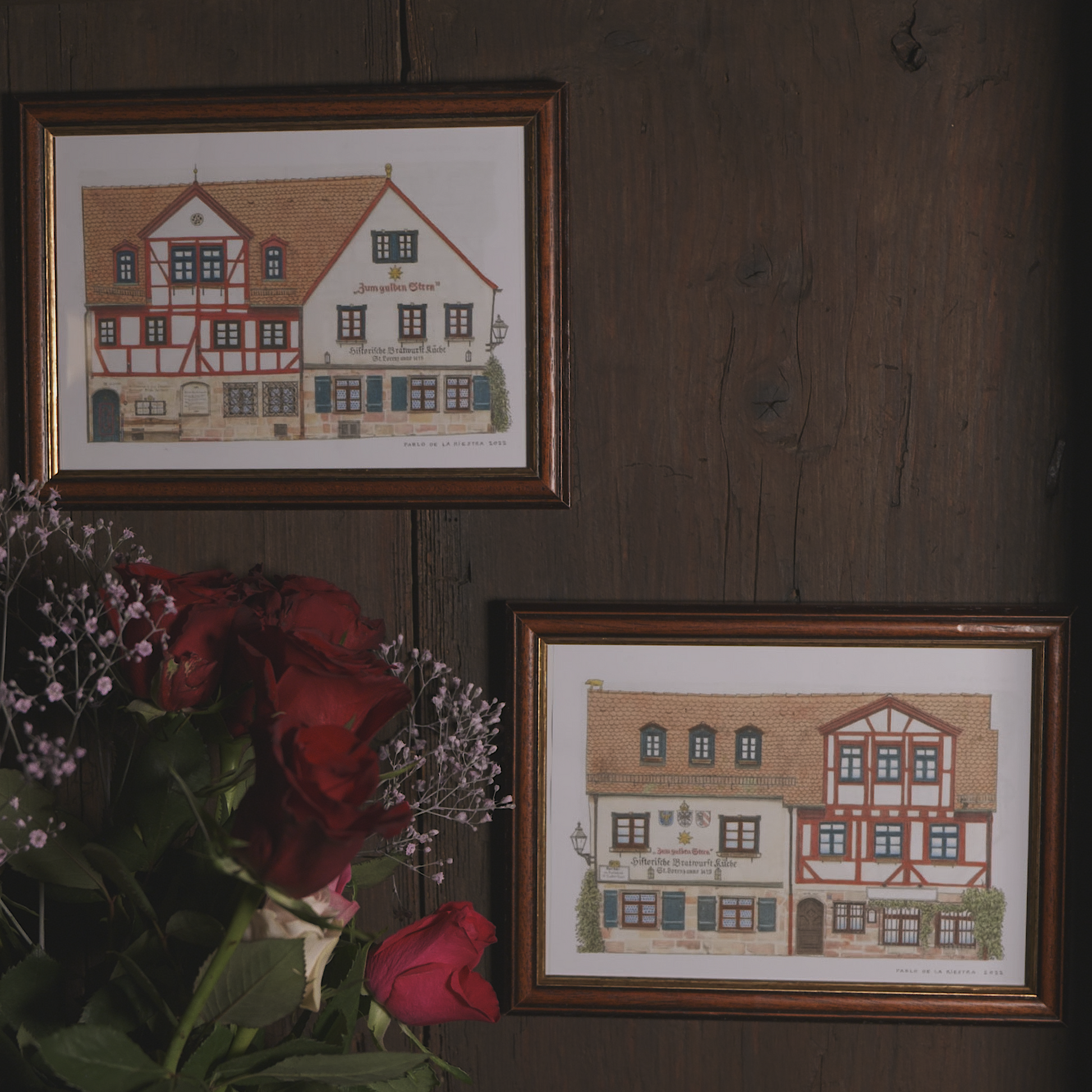 Zwei gerahmte Kunstdrucke mit Illustrationen der historischen Bratwurstküche „Zum Gulden Stern“ vom Künstler Pablo de la Riestra, aufgehängt an einer dunklen Holzwand. Im Vordergrund liegen rote Rosen und zarte Blumenarrangements.