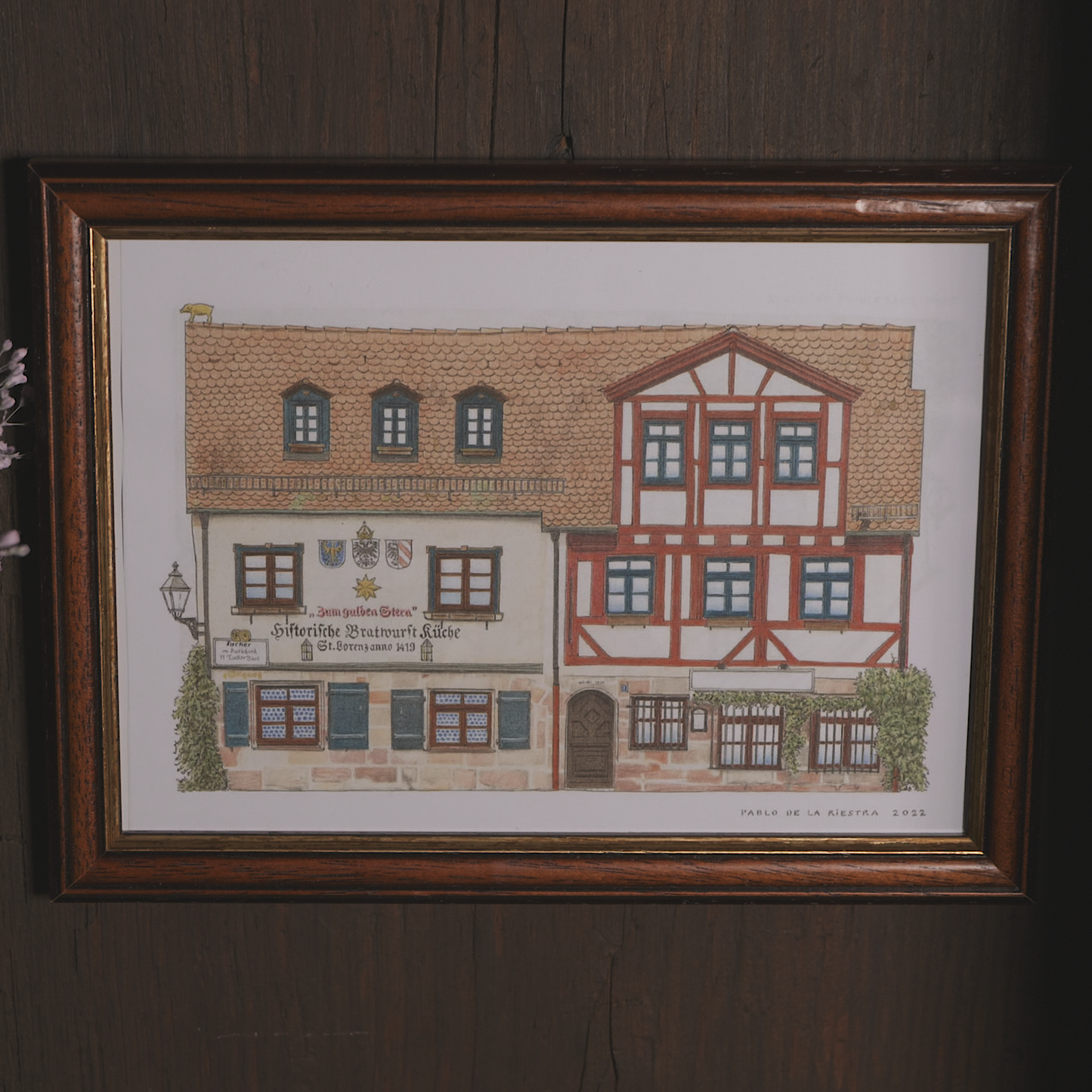 Gerahmter Kunstdruck der historischen Bratwurstküche „Zum Gulden Stern“ in Nürnberg, gezeichnet vom Künstler Pablo de la Riestra. Die Illustration zeigt die Fachwerkarchitektur des Gebäudes mit roten Balken, kleinen Fenstern und historischen Wappen an der Fassade. Der Rahmen aus dunklem Holz hängt an einer rustikalen Holzwand.