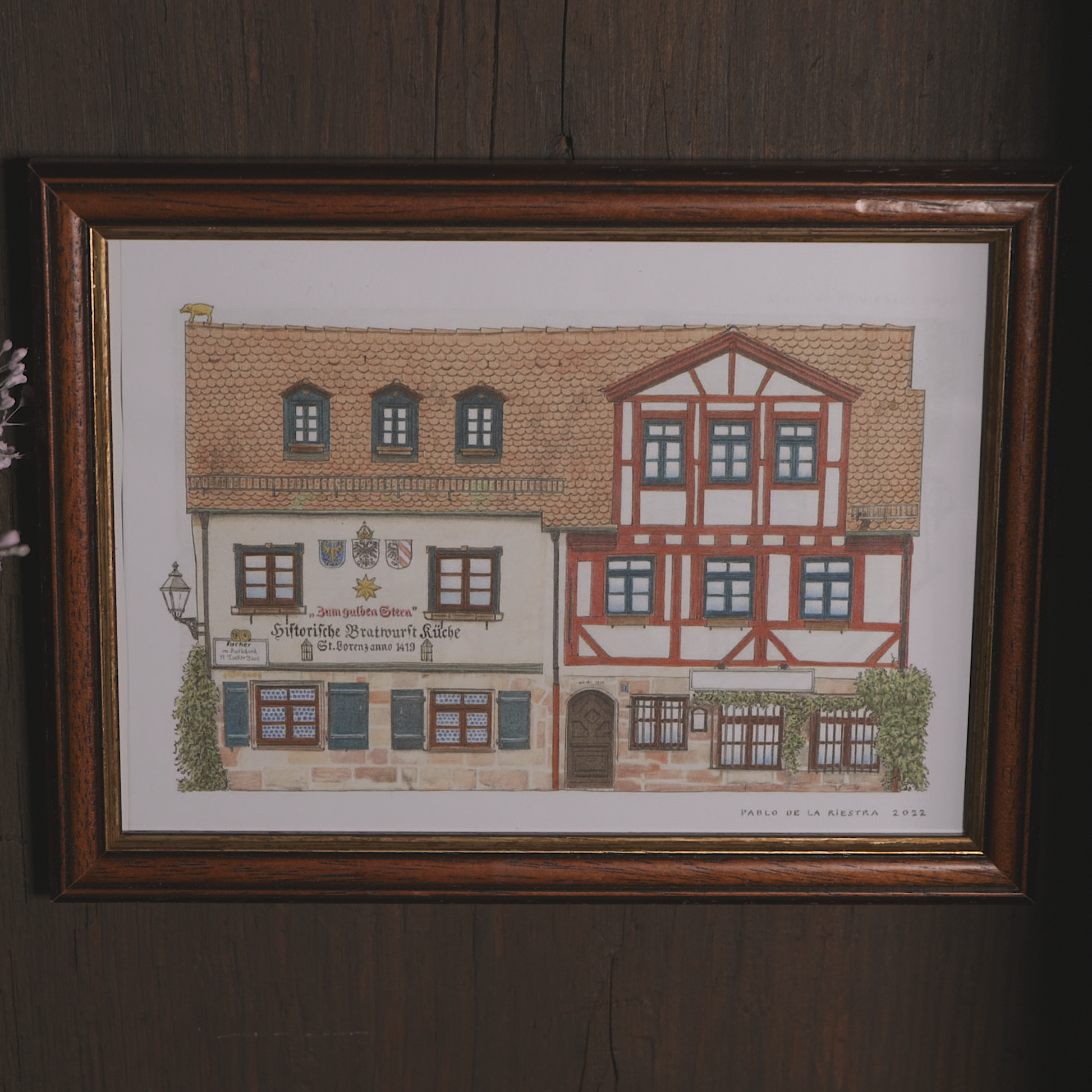 Gerahmter Kunstdruck der historischen Bratwurstküche „Zum Gulden Stern“ in Nürnberg, gezeichnet vom Künstler Pablo de la Riestra. Die Illustration zeigt die Fachwerkarchitektur des Gebäudes mit roten Balken, kleinen Fenstern und historischen Wappen an der Fassade. Der Rahmen aus dunklem Holz hängt an einer rustikalen Holzwand.