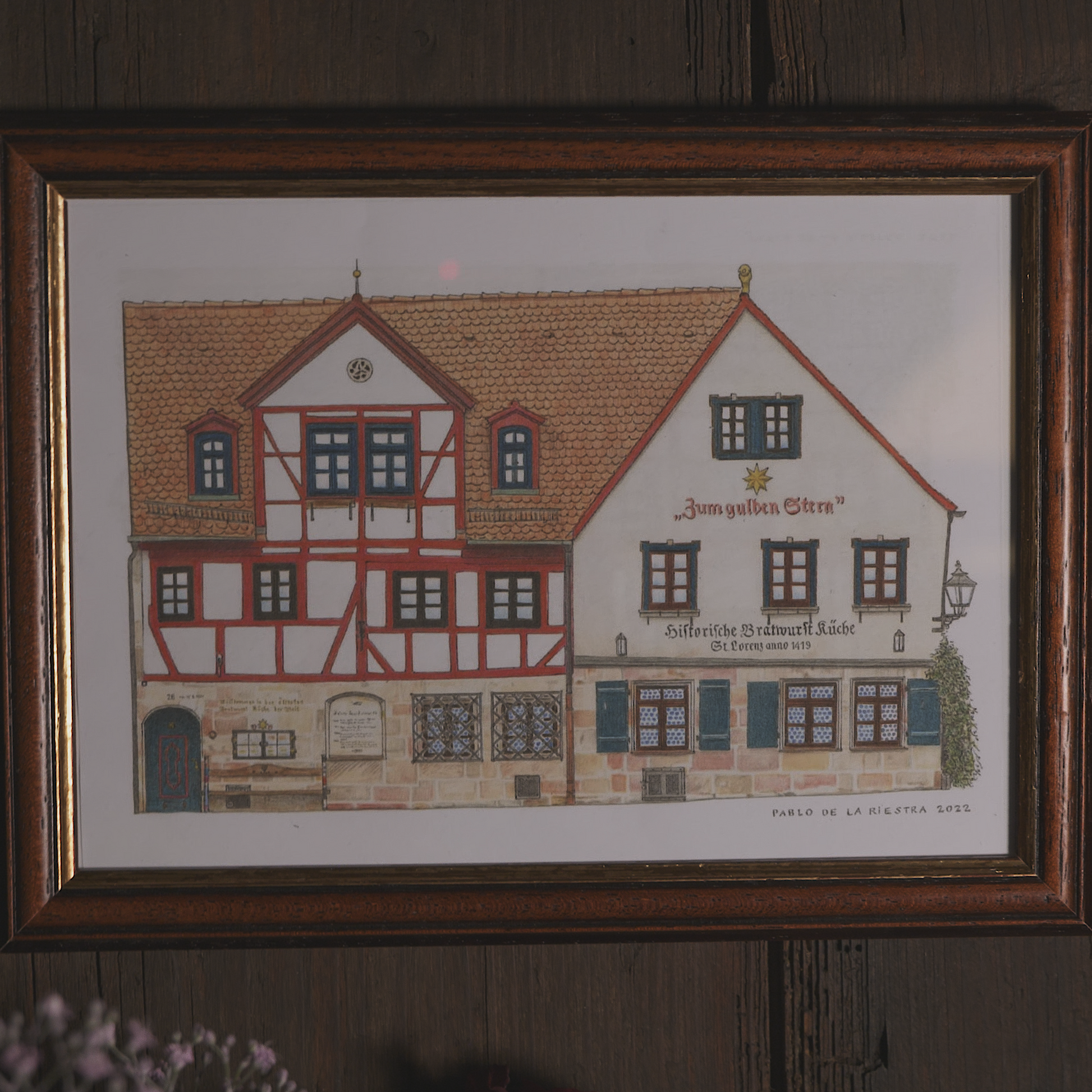 Detailreicher Kunstdruck des historischen Fachwerkgebäudes „Zum Gulden Stern“ in Nürnberg, dem ältesten Bratwurstrestaurant der Welt. Das Werk stammt vom Künstler Pablo de la Riestra und zeigt die charakteristische Fachwerkfassade mit roten Balken, kleinen Fenstern und der historischen Beschriftung. Der Druck ist in einem dunkelbraunen Holzrahmen gefasst und hängt an einer rustikalen Holzwand.