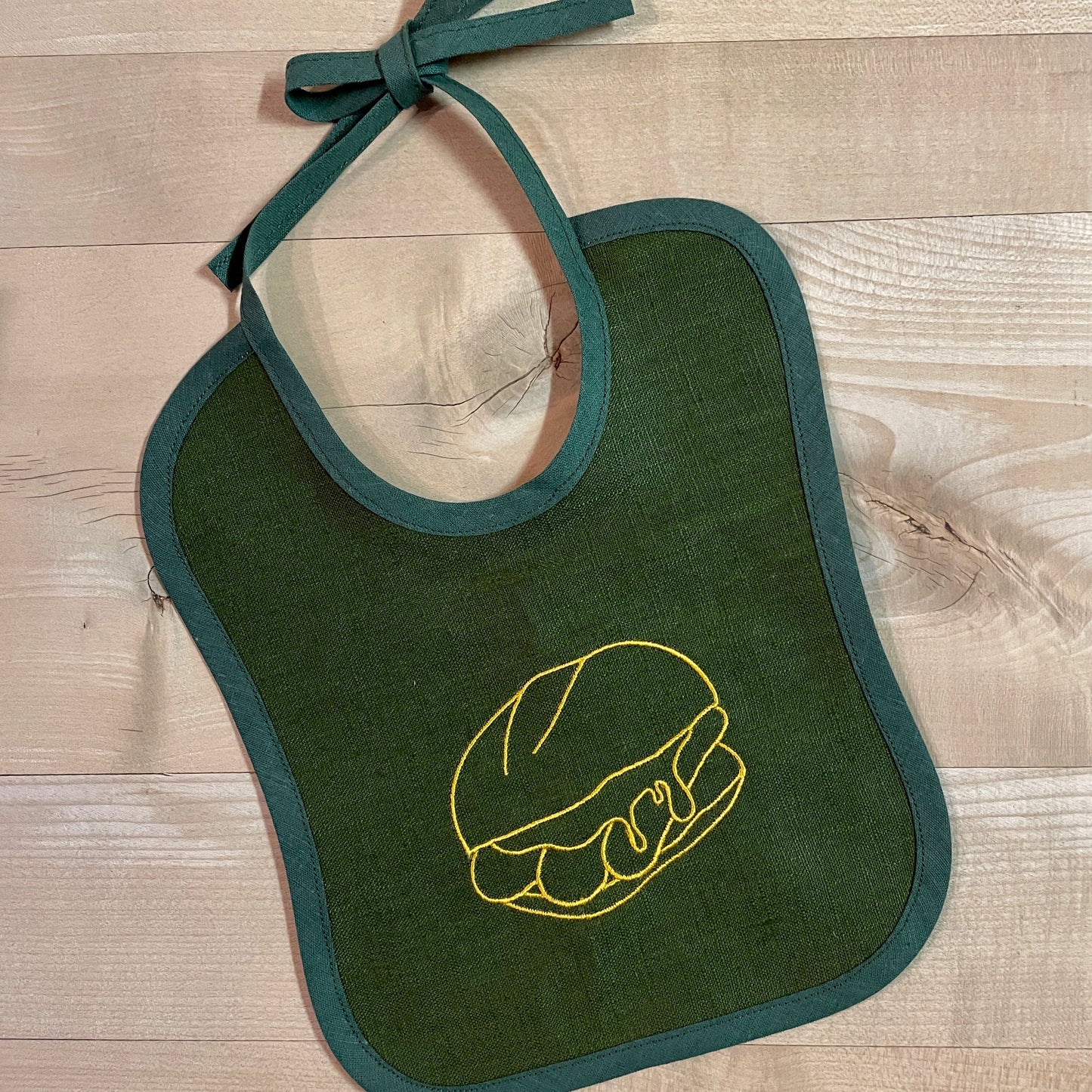 Ein dunkelgrünes Baby-Lätzchen aus Leinen mit einer goldgelben Stickerei eines Bratwurstbrötchens ("3 im Weggla") liegt auf einem hellen Holztisch. Das Lätzchen hat eine dunkelgrüne Einfassung und lange Stoffbänder zum Binden am oberen Rand. Die feine Stickerei hebt das Motiv stilvoll hervor und verleiht dem Lätzchen eine besondere, traditionelle Note.