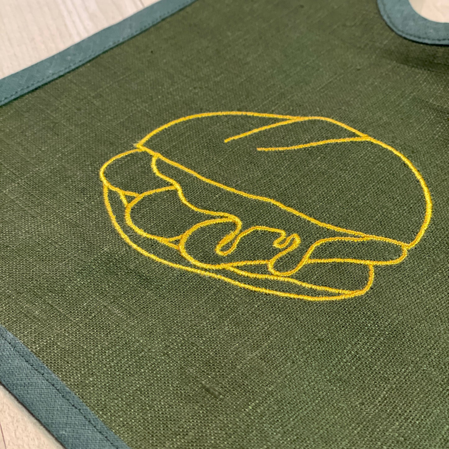 Nahaufnahme eines dunkelgrünen Baby-Lätzchens aus Leinen mit einer goldgelben Stickerei eines Bratwurstbrötchens ("3 im Weggla"). Die feine Stickerei hebt das Motiv präzise hervor und verleiht dem Stoff eine edle Optik. Der Rand des Lätzchens ist mit einem dunkelgrünen Stoff eingefasst. Die Textur des Leinens ist gut sichtbar und unterstreicht die hochwertige Verarbeitung des Lätzchens.